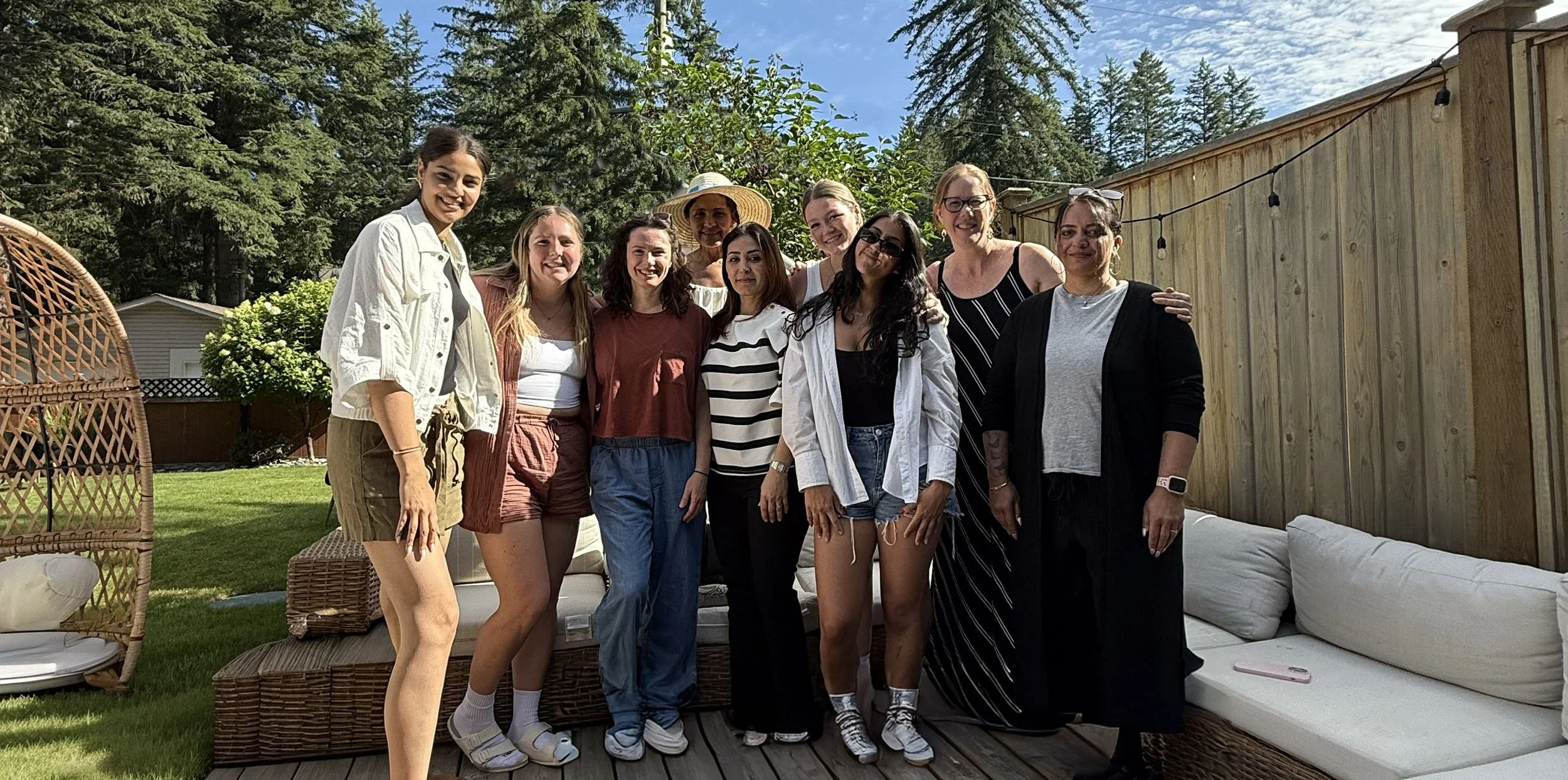 A group of nine Twig + Owl female educators standing together outdoors on a sunny day, smiling for a photo, with trees, a wooden fence, and a sky with clouds in the background.