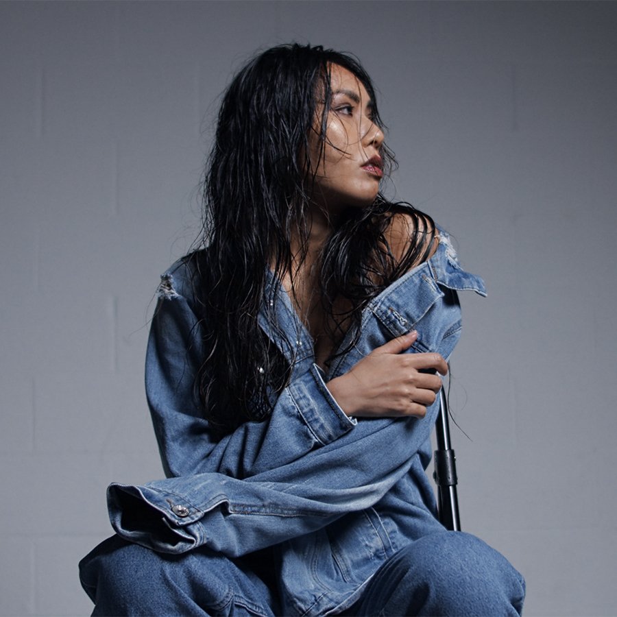 A woman with wet dark hair wearing an oversized denim jacket sitting against a plain gray wall.