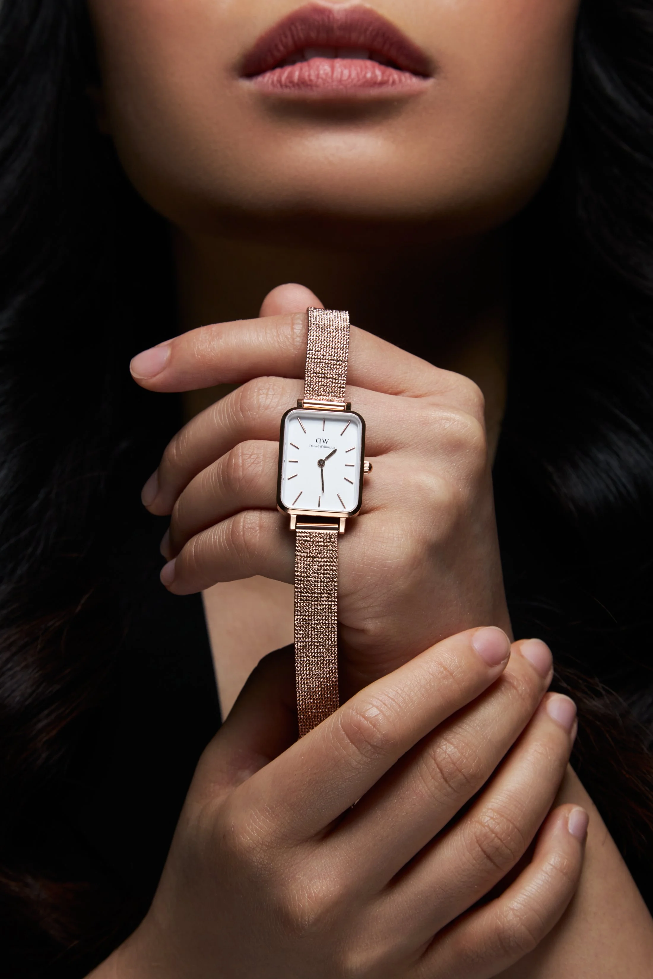 A woman with dark hair is holding a rectangular watch with a white face and gold band near her face.