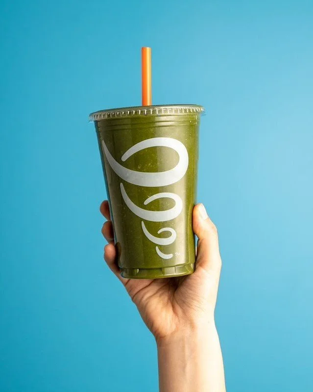A hand holding a green smoothie in a cup with a clear plastic lid and an orange straw, with a blue background.