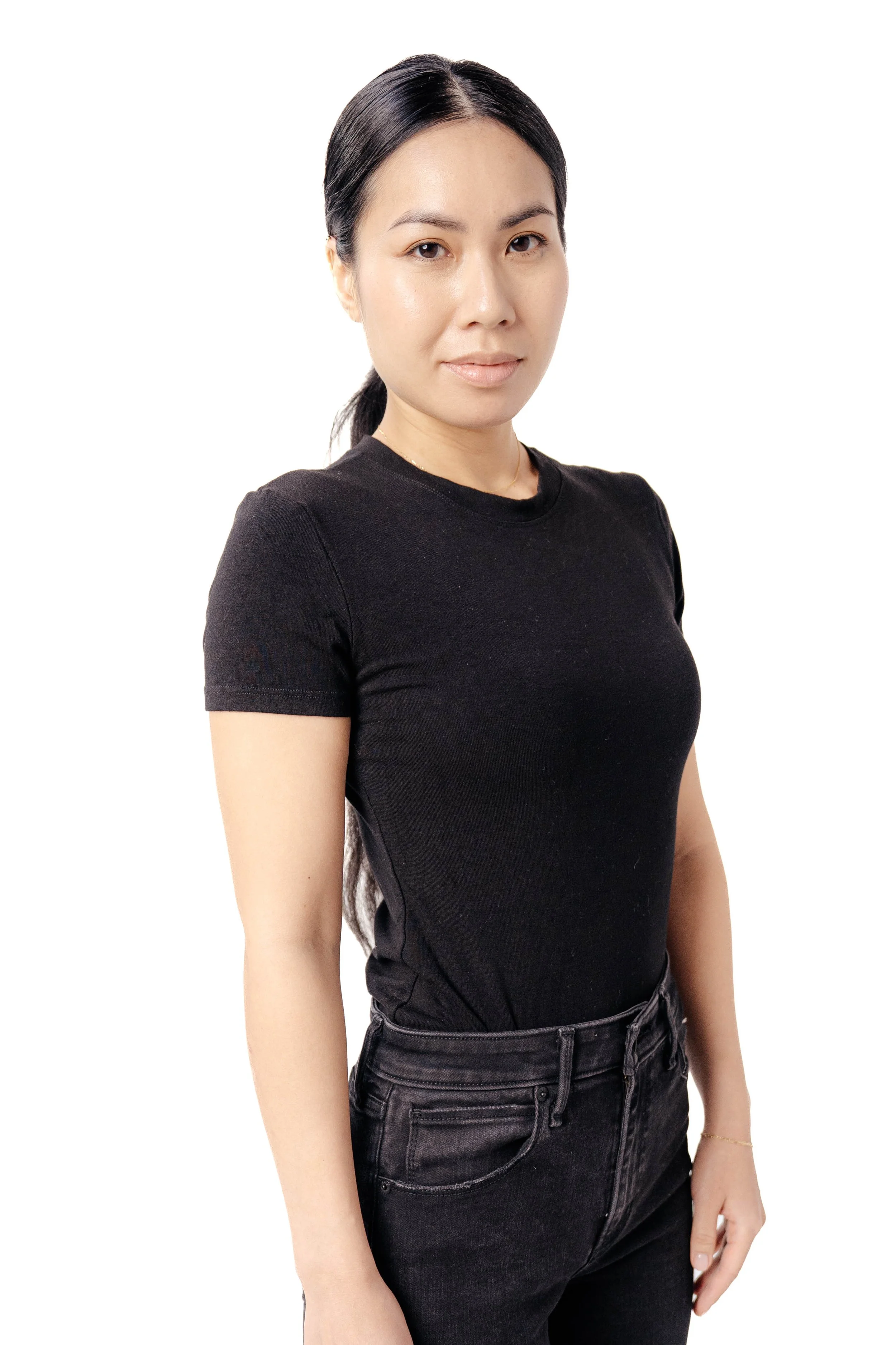 A woman with long black hair wearing a black t-shirt and black jeans standing against a white background.