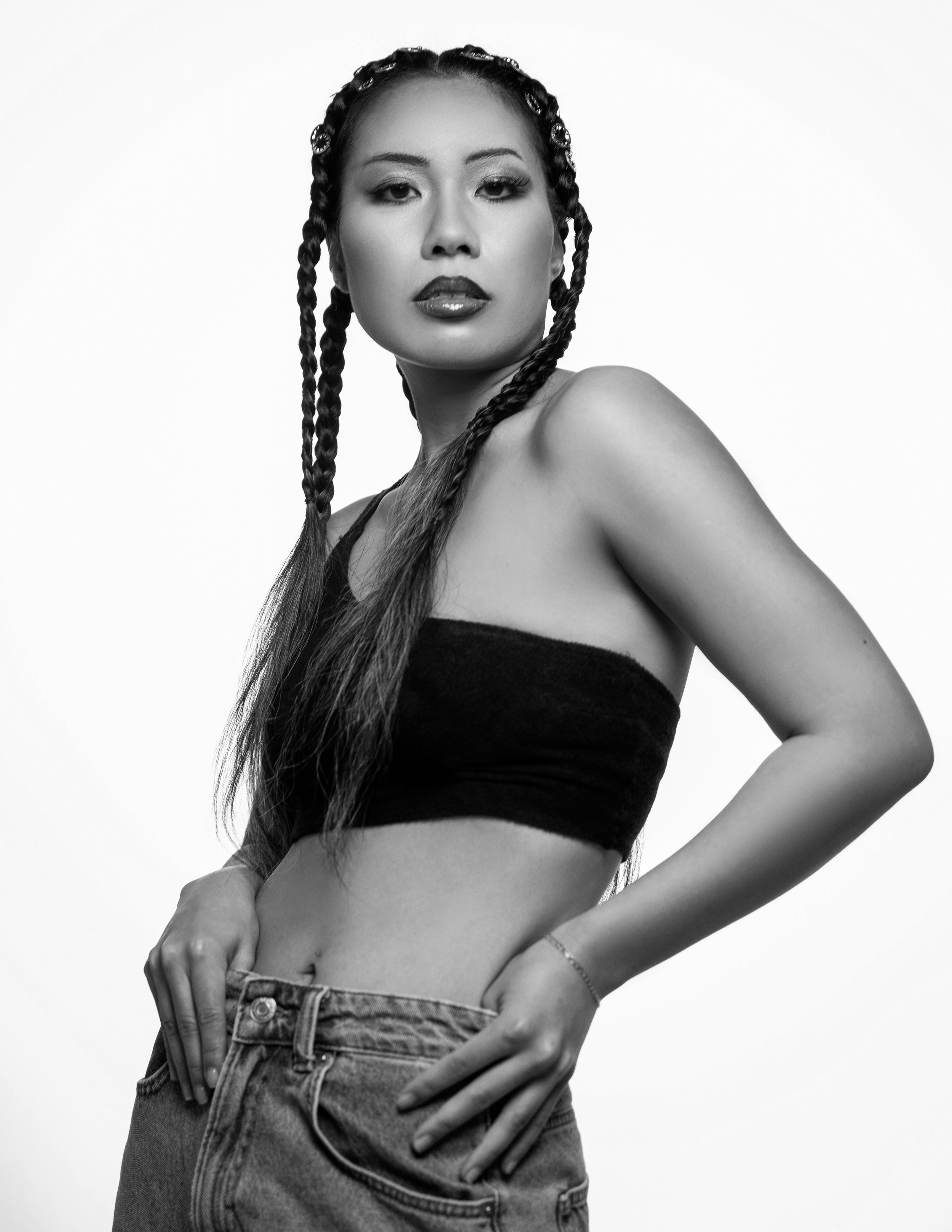 Black and white photo of a young woman with braided hair, wearing a strapless top and jeans, posing with her hands on her hips against a plain background.