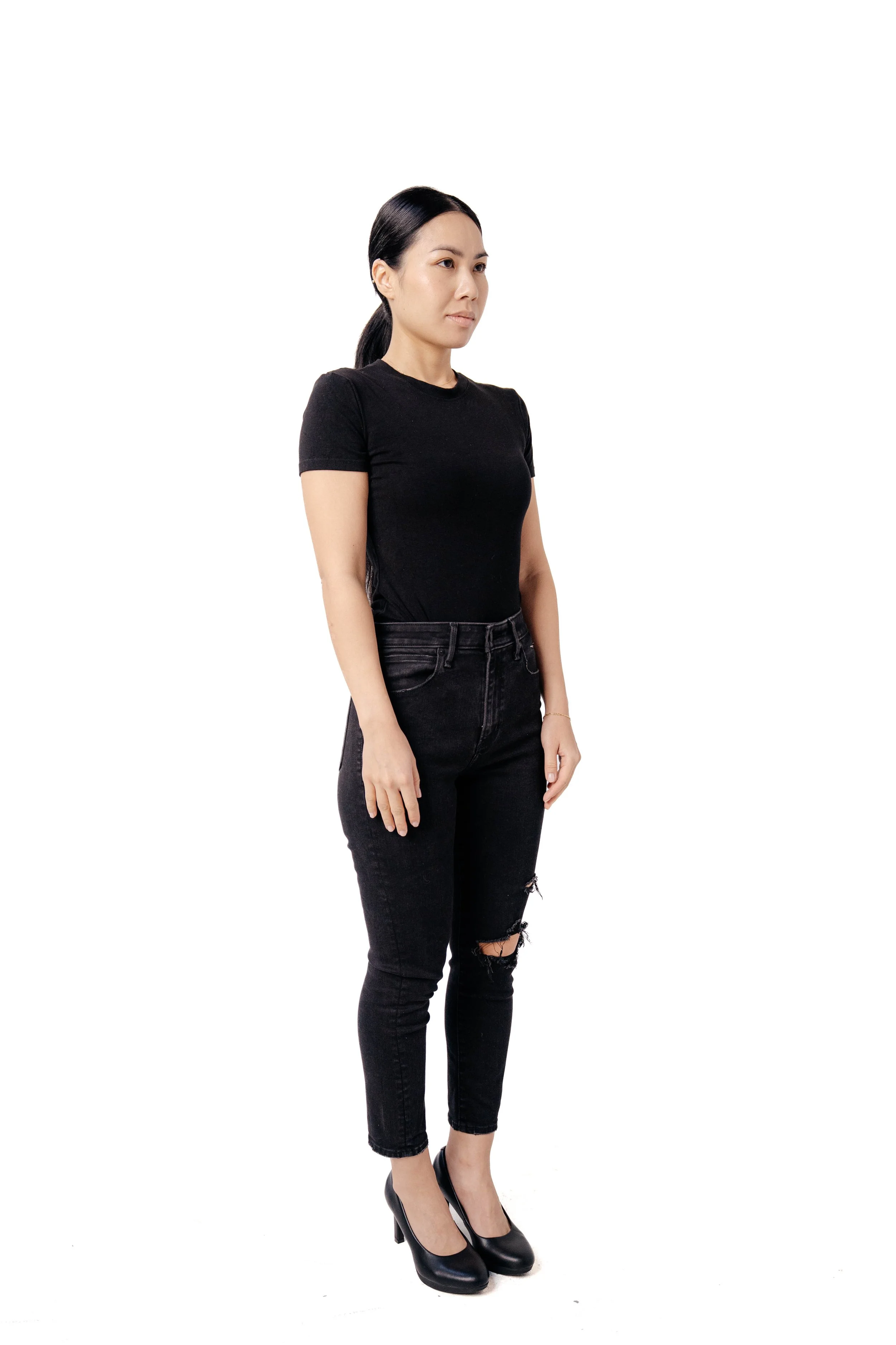 A woman with long dark hair in a ponytail, wearing a black t-shirt and black ripped jeans, standing against a white background.