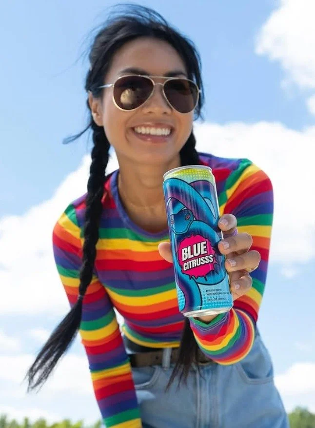 A woman with long dark hair in braids wearing sunglasses and a colorful rainbow striped shirt, smiling and holding a can of Blue Citrus soda outdoors under a partly cloudy sky.