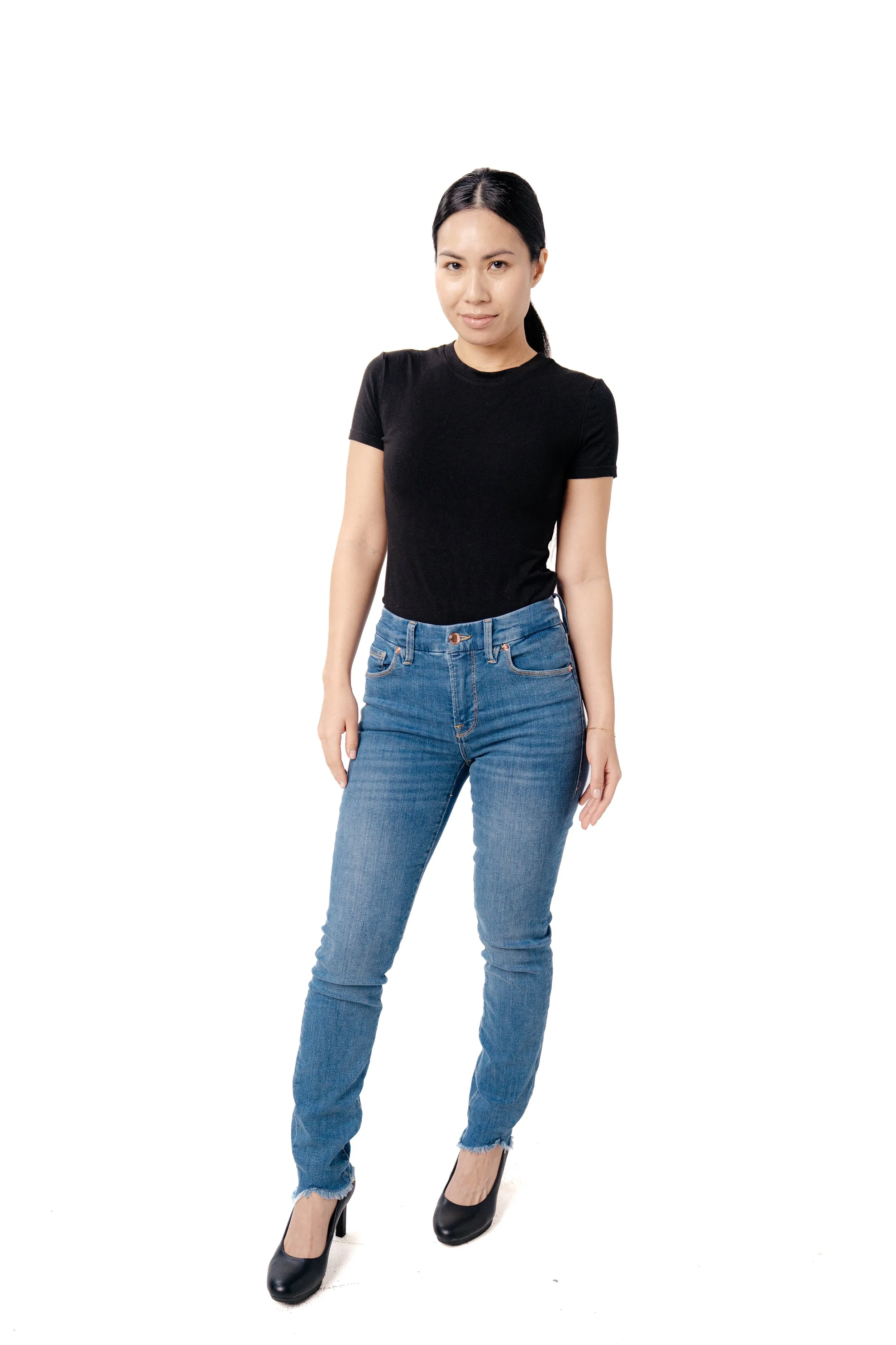 A woman with black hair in a ponytail, wearing a black t-shirt, blue jeans, and black heels, standing with one leg slightly bent on a white background.
