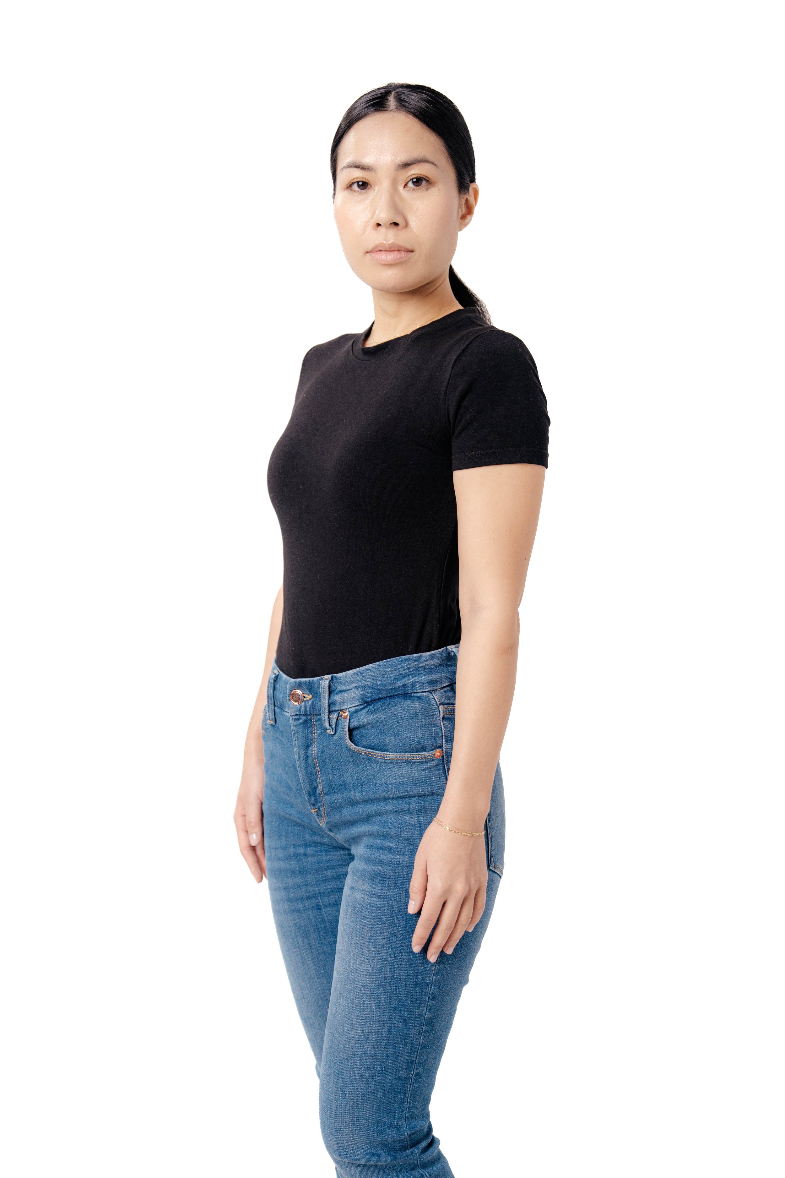 A woman standing against a white background, wearing a black T-shirt and blue jeans, with her hair tied back.