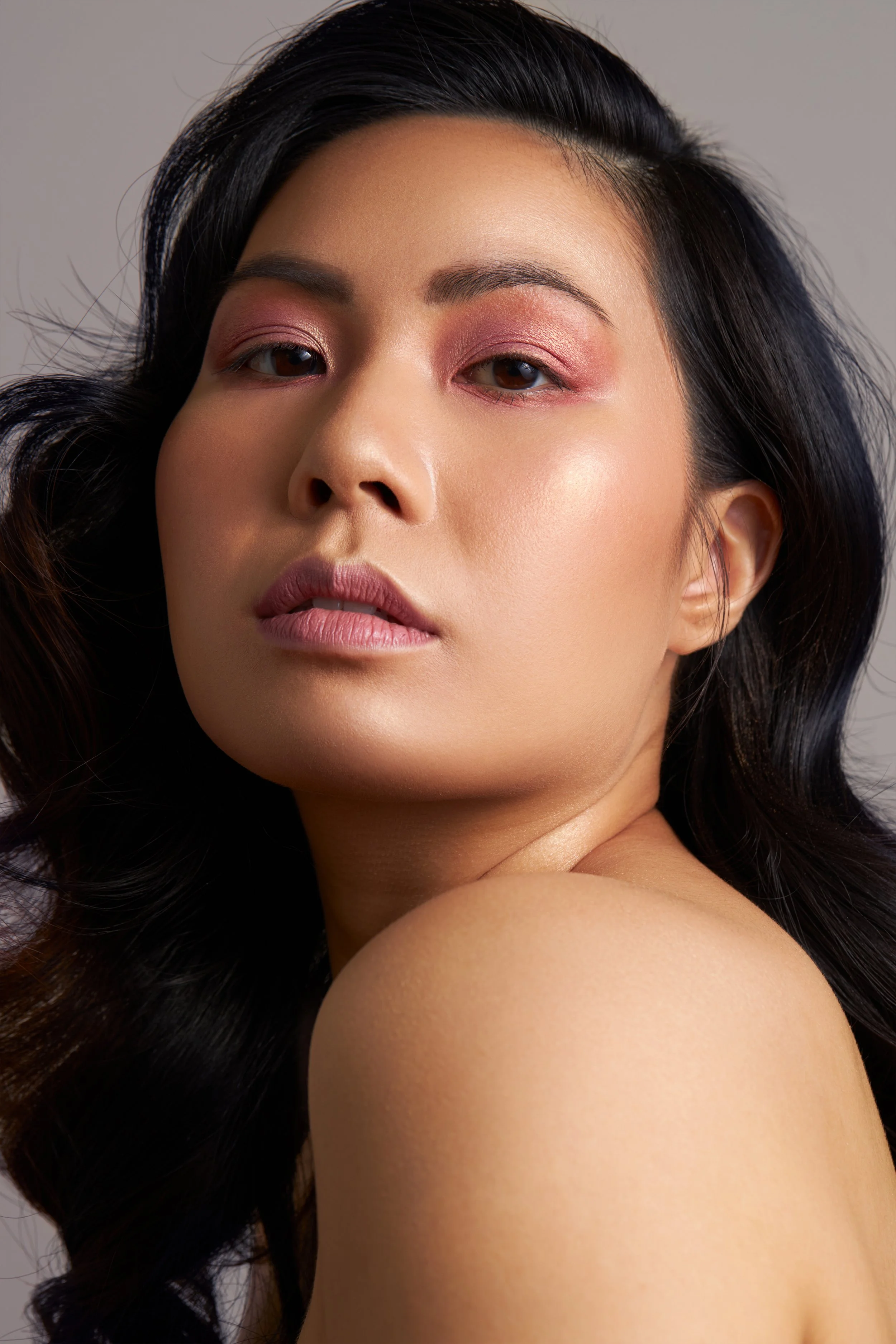 A close-up portrait of a woman with long, dark, wavy hair and makeup with pink eyeshadow and soft pink lipstick.