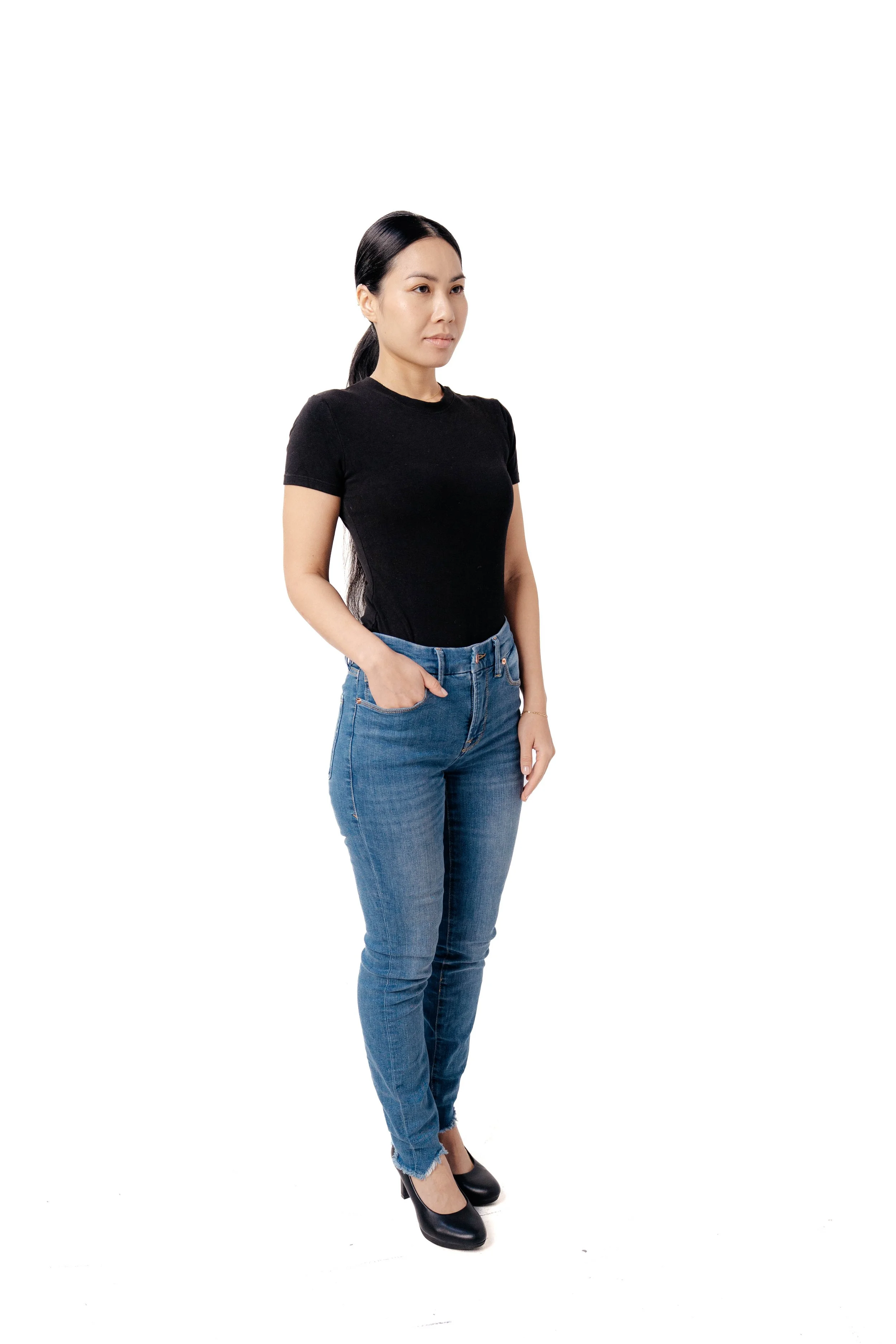 A woman with long black hair wearing a black t-shirt and blue jeans standing with one hand in her pocket against a plain white background.
