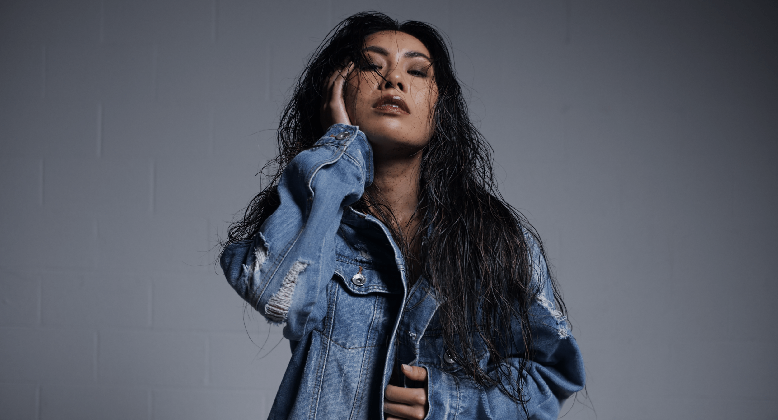A woman with long, wet, curly dark hair wearing a distressed denim jacket, posing against a plain gray wall.