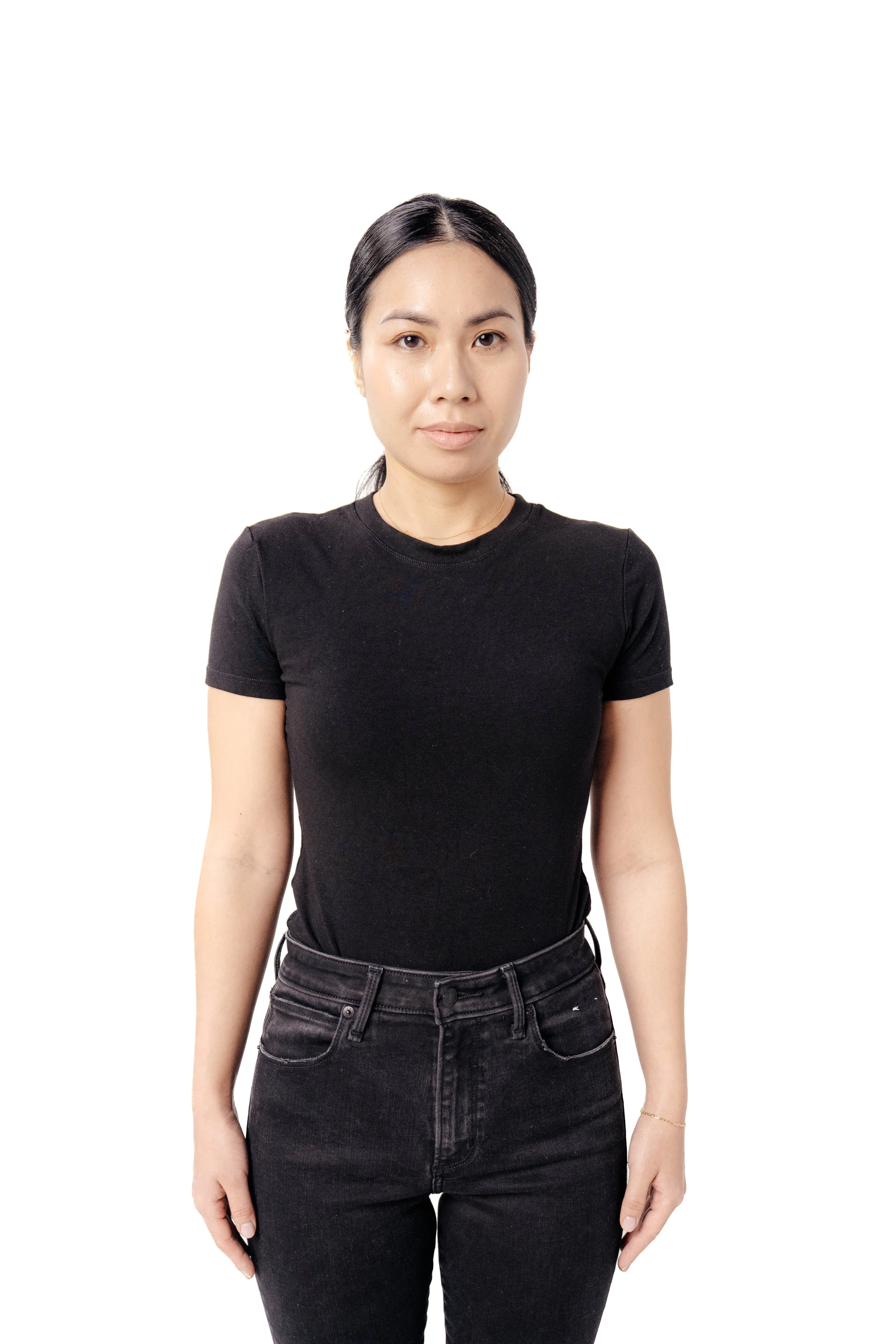 A woman with black hair tied back, wearing a black t-shirt and black jeans, standing against a white background.