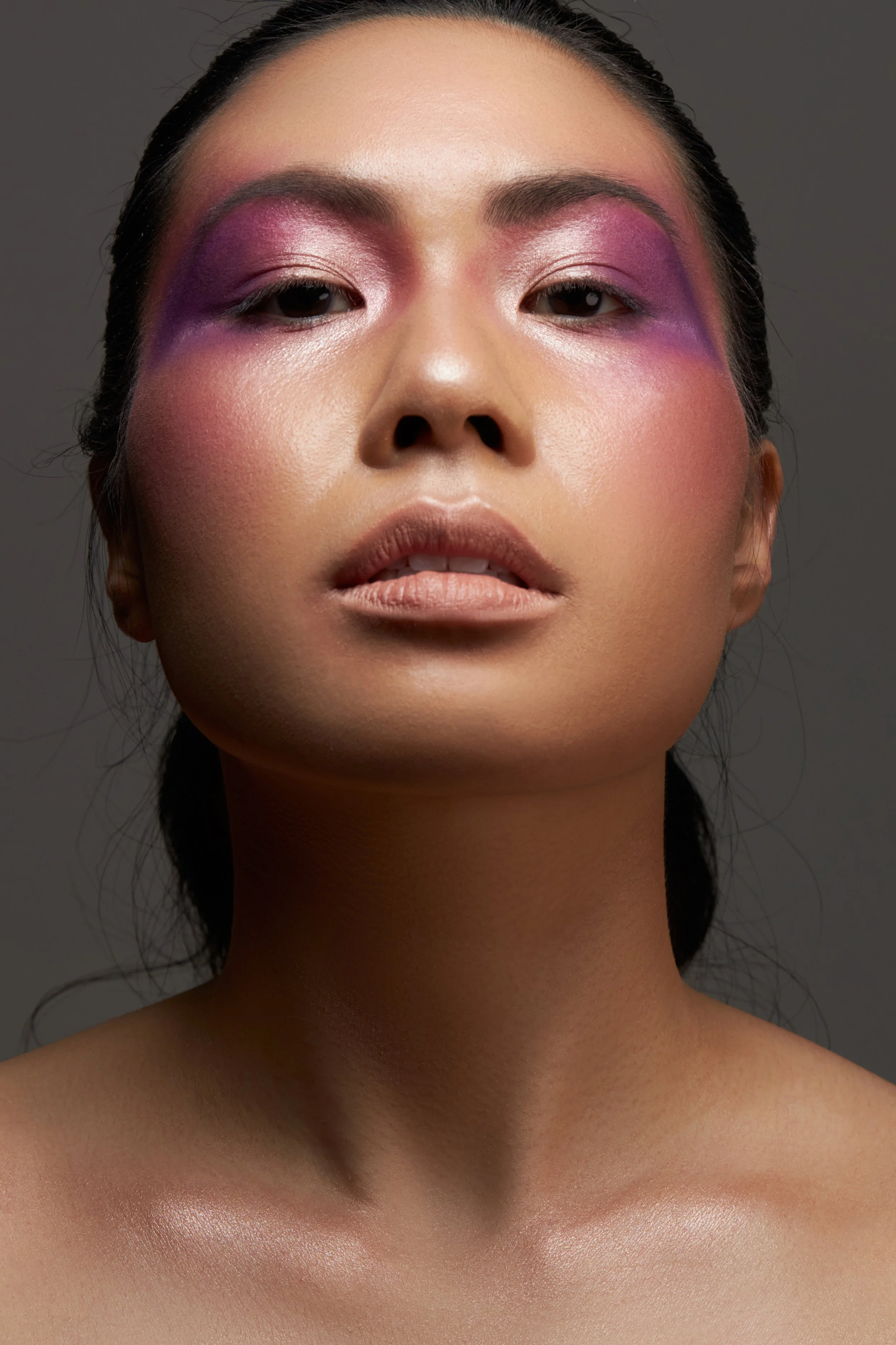 Close-up of a woman with pink and purple eyeshadow, nude lipstick, and a smooth complexion, looking directly at the camera against a gray background.
