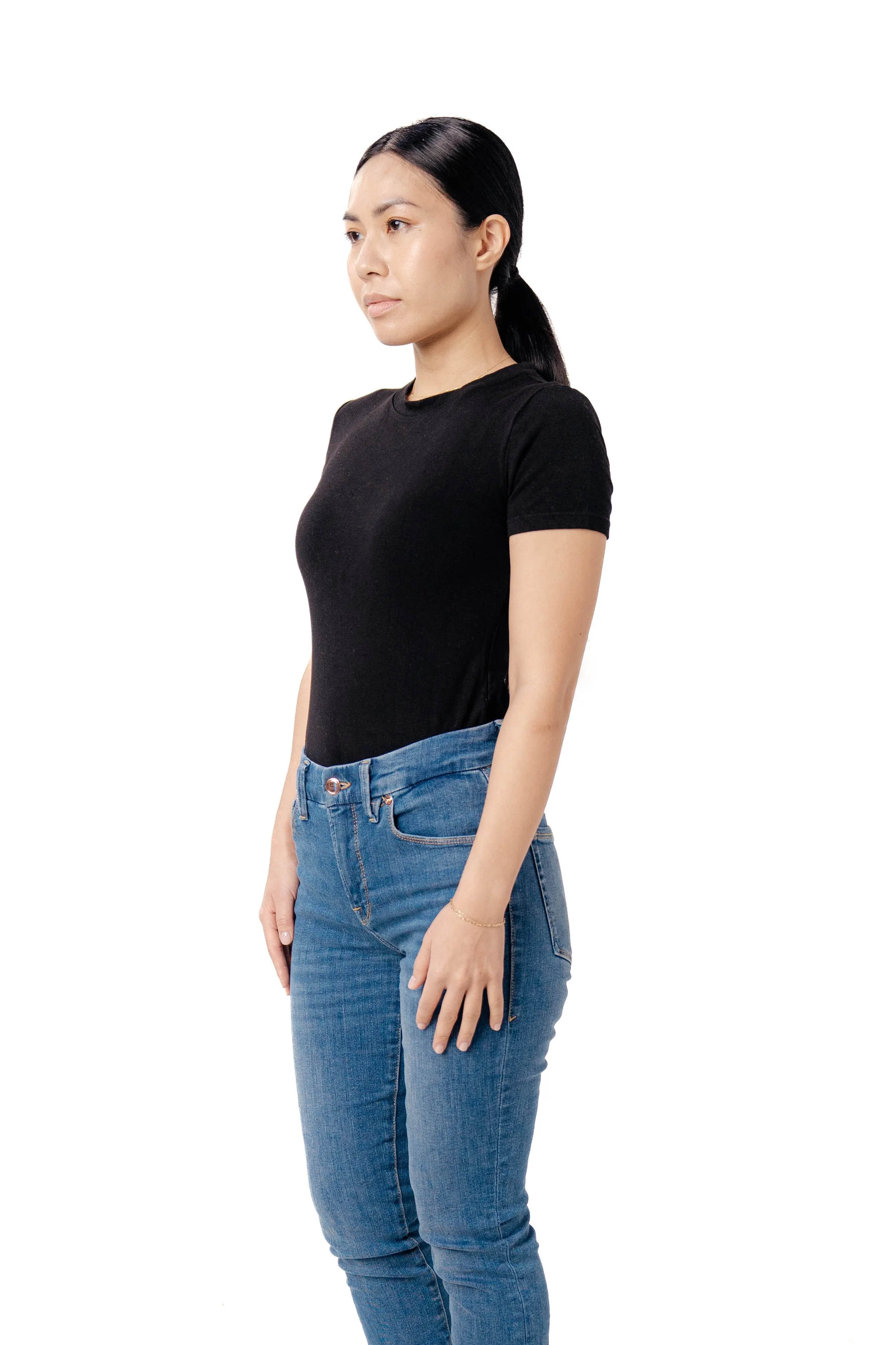 Young woman with dark hair tied back, wearing a black t-shirt and blue jeans, standing against a white background.