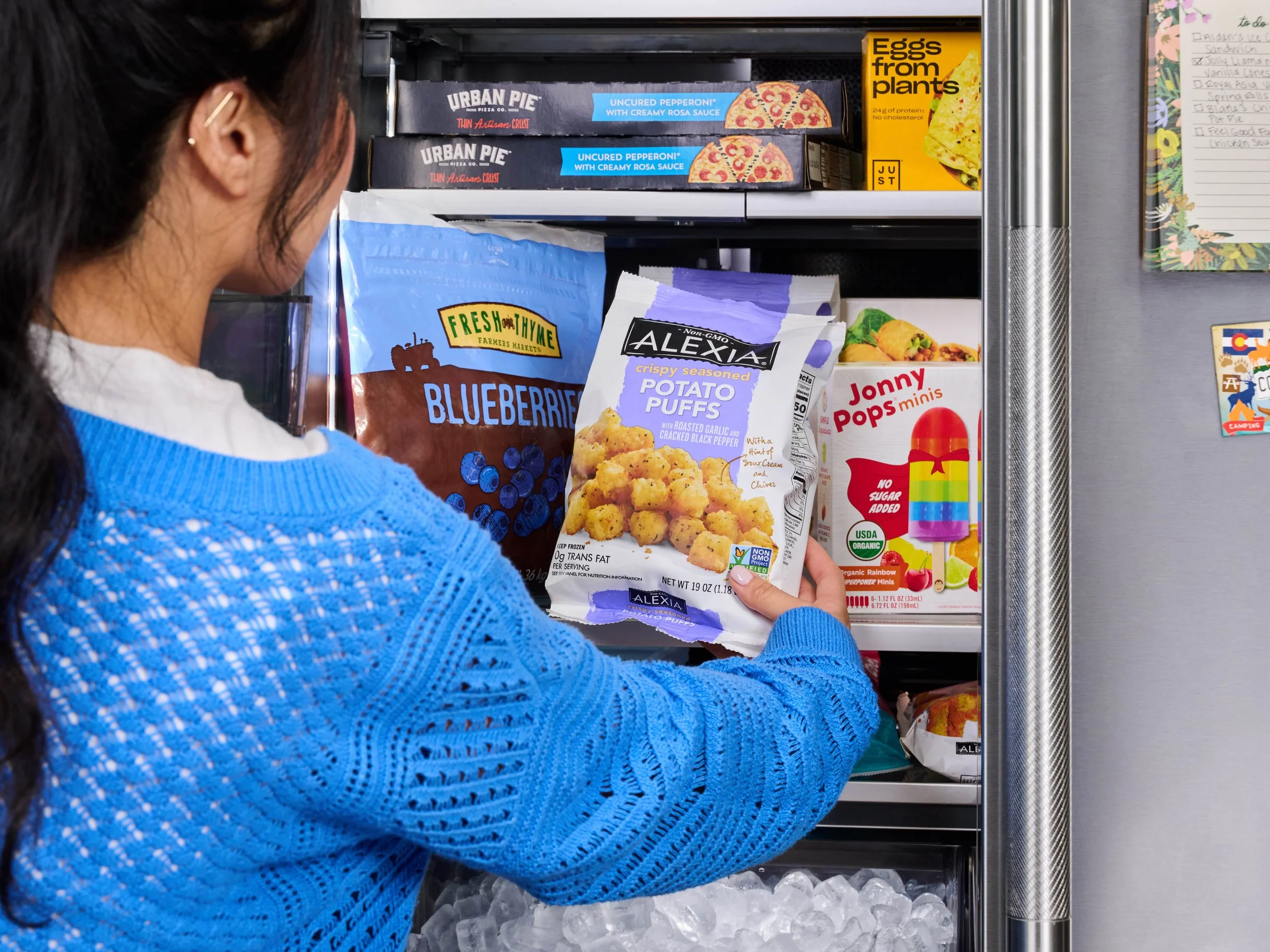 Woman in a blue sweater selecting Alexia crispy seasoned potato puffs from a freezer shelf.