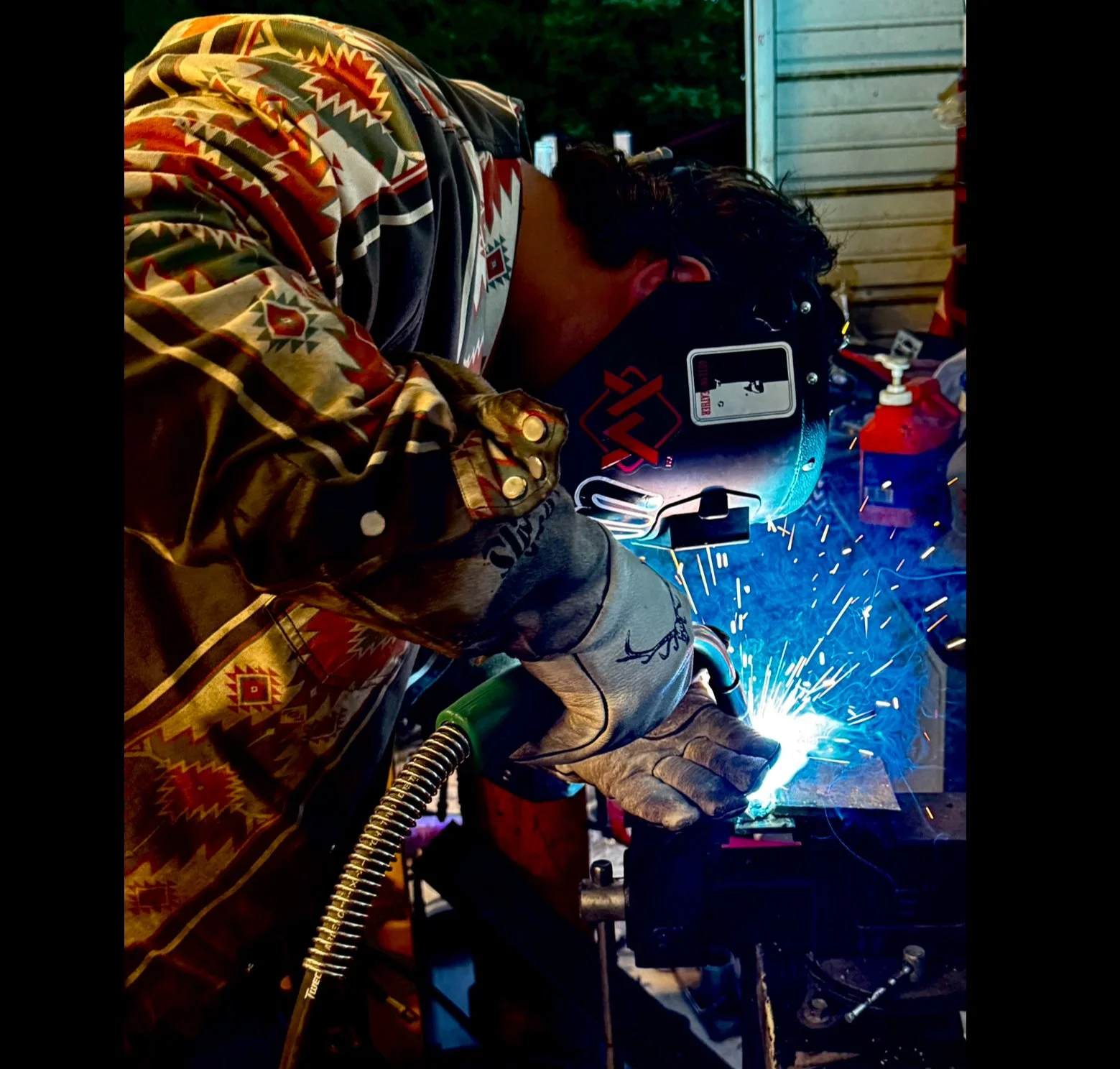 A person welding metal outdoors at night, wearing a patterned shirt and a welding helmet, with sparks flying from the welding process.