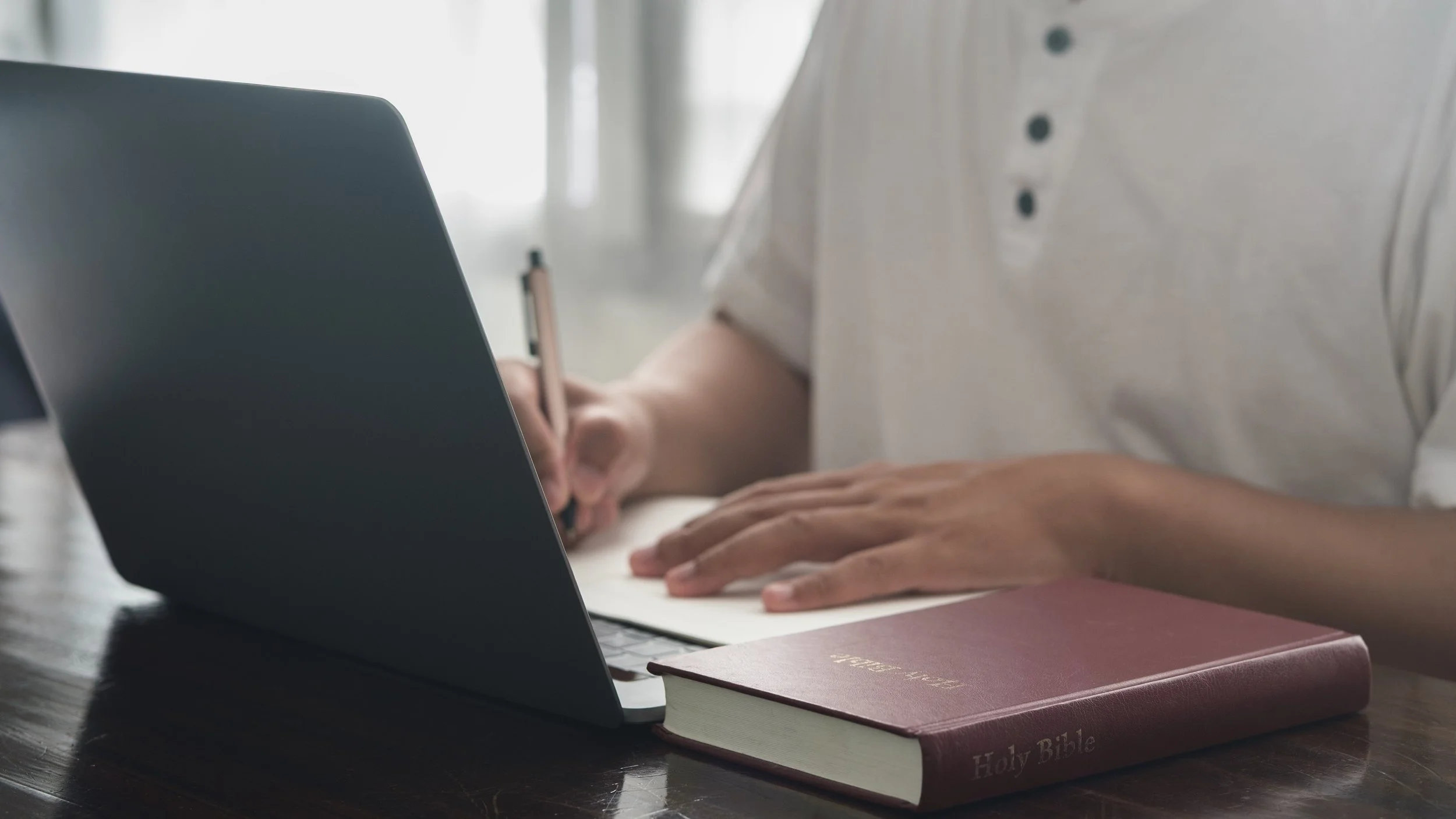A person working at a desk with a laptop, a Bible, and a notebook with a pen, with sunlight coming through a window in the background.