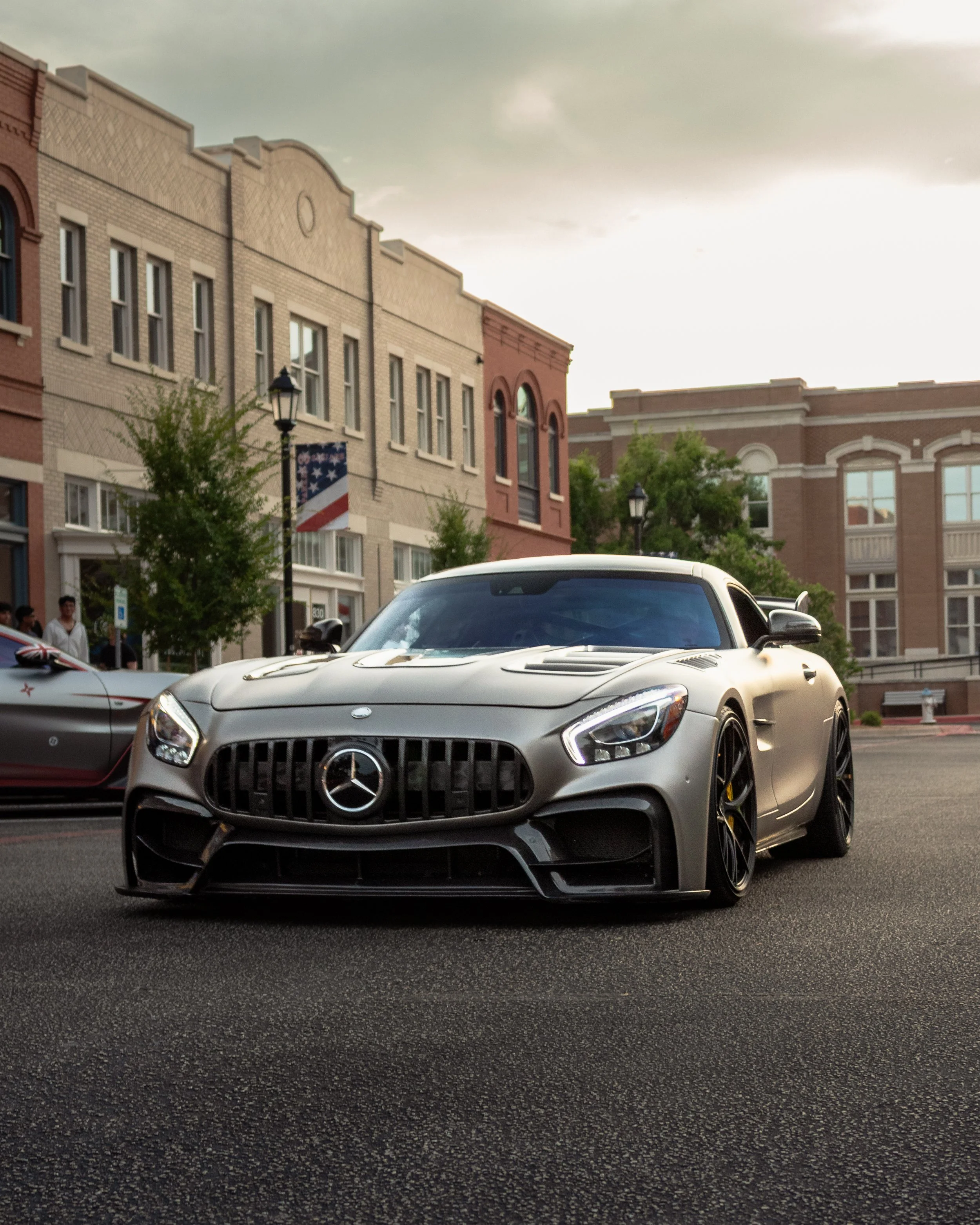 A silver Mercedes-Benz AMG GT sports car parked on a city street with brick buildings and trees in the background.