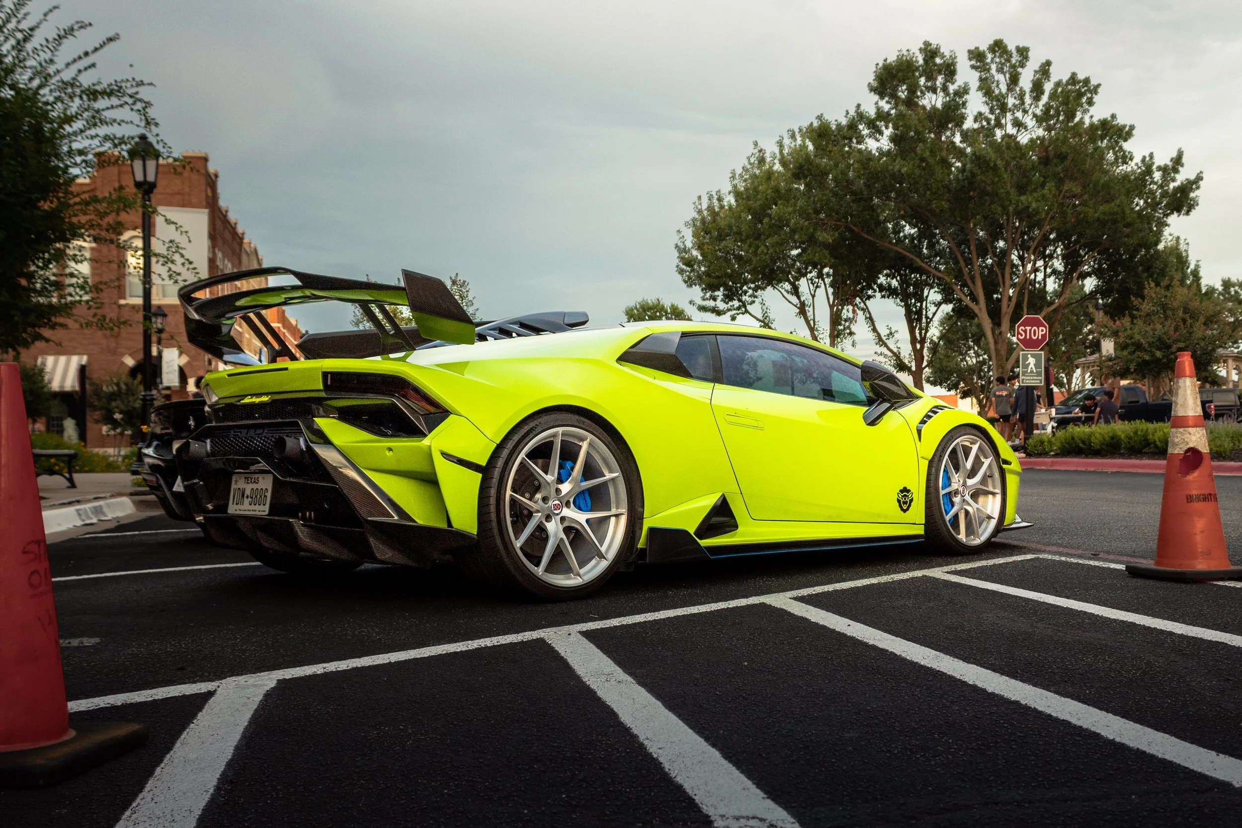 A neon yellow Lamborghini supercar parked in a marked parking space, surrounded by safety cones, with trees, a stop sign, and people in the background.
