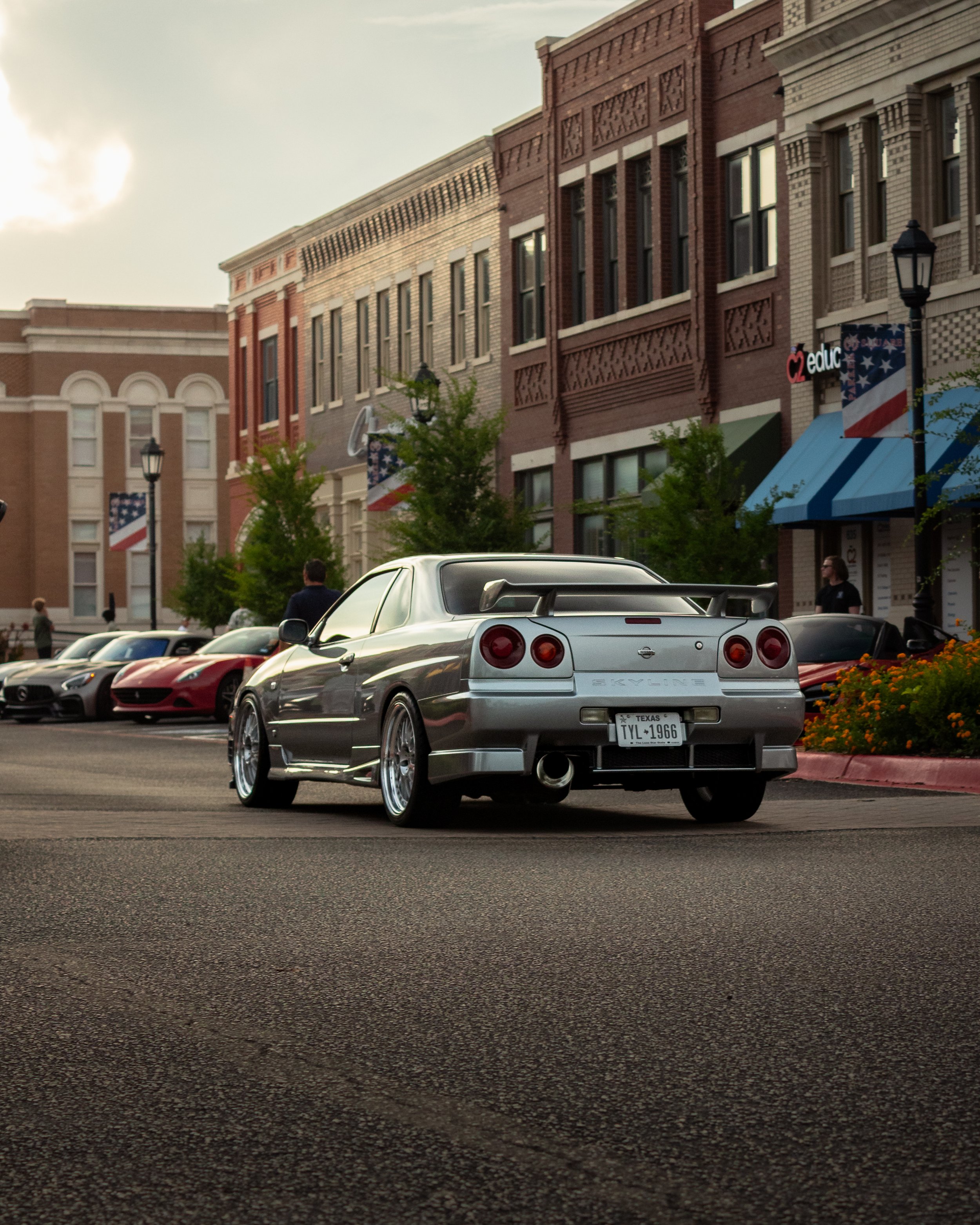 A silver Nissan Skyline car with a large rear spoiler parked on a city street in front of a row of buildings with American flags and streetlights.