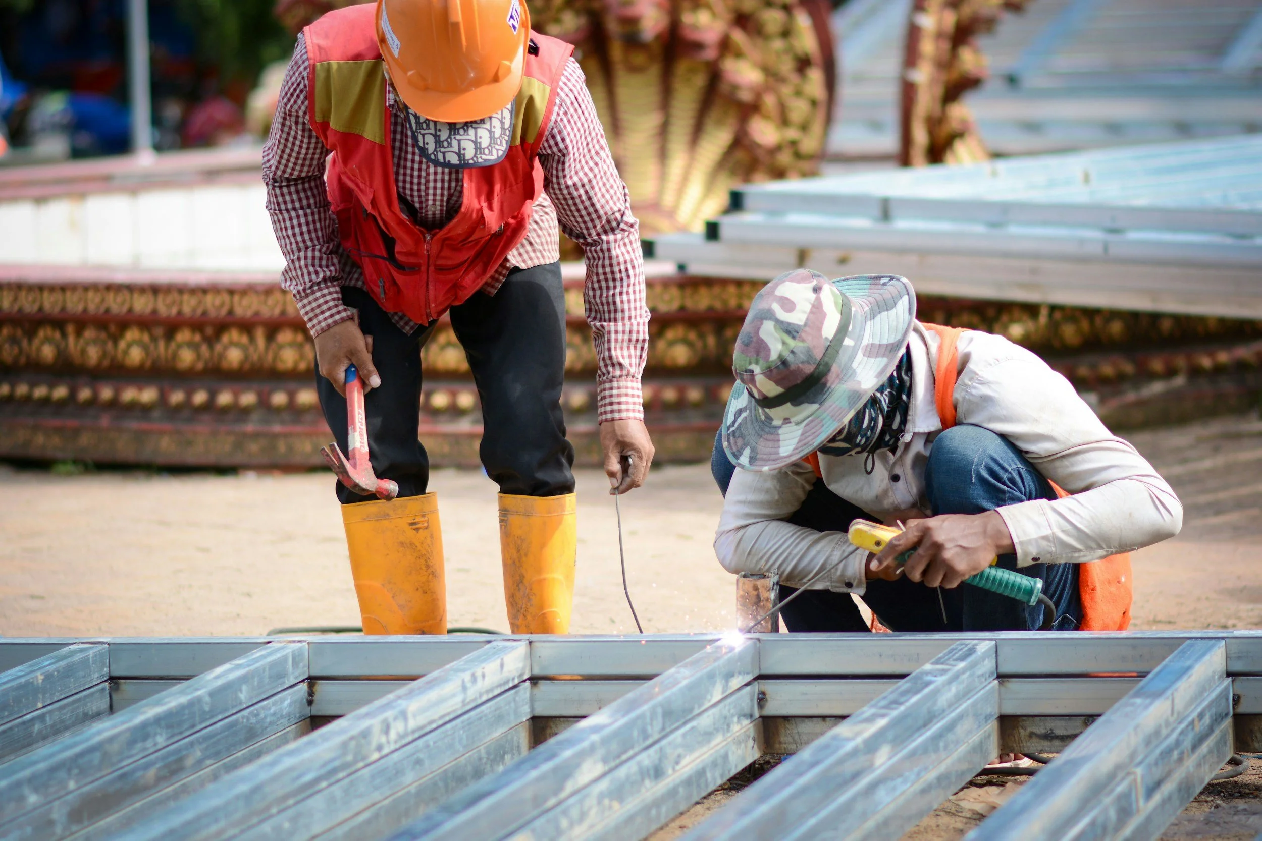 Two construction workers wearing safety hats and vests working on metal framing at a construction site.