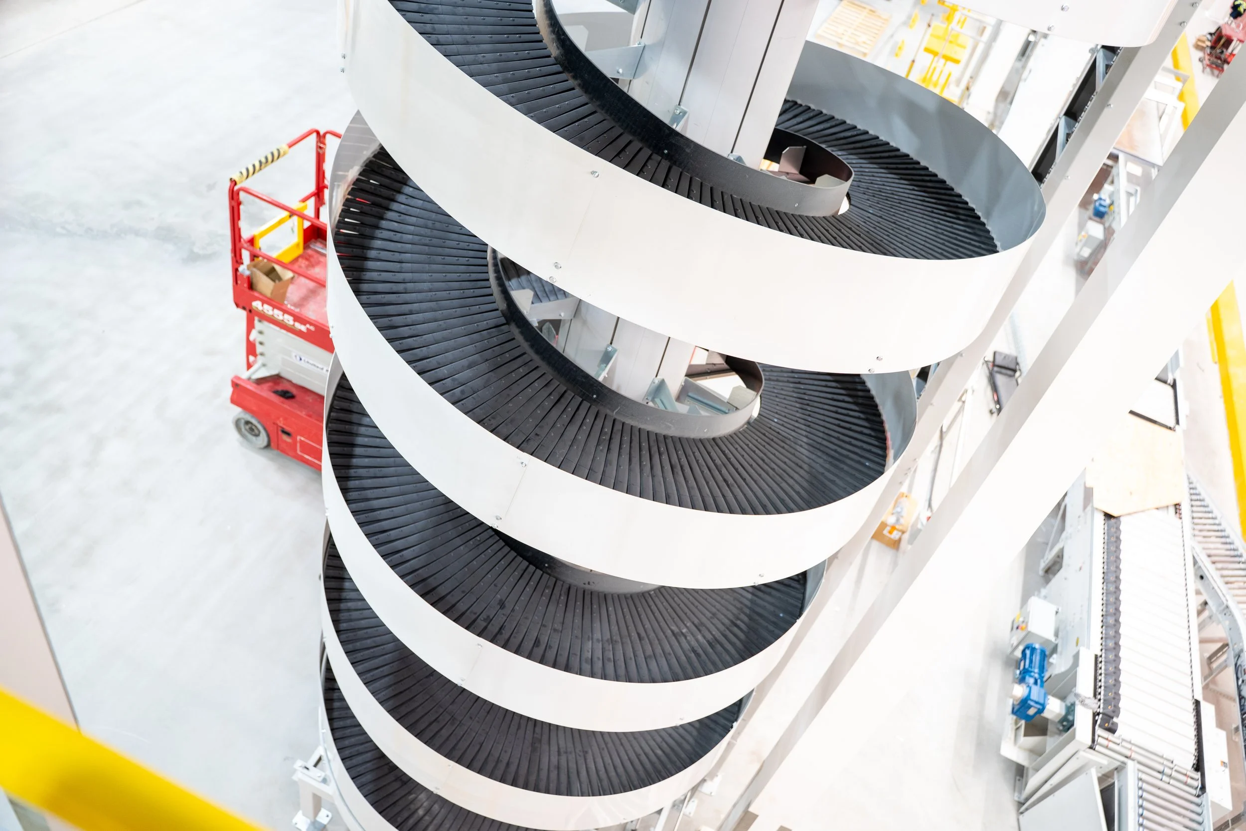 Aerial view of a vertical spiral conveyor with black rollers and white side panels, situated in an industrial warehouse with equipment and a red scissor lift nearby.