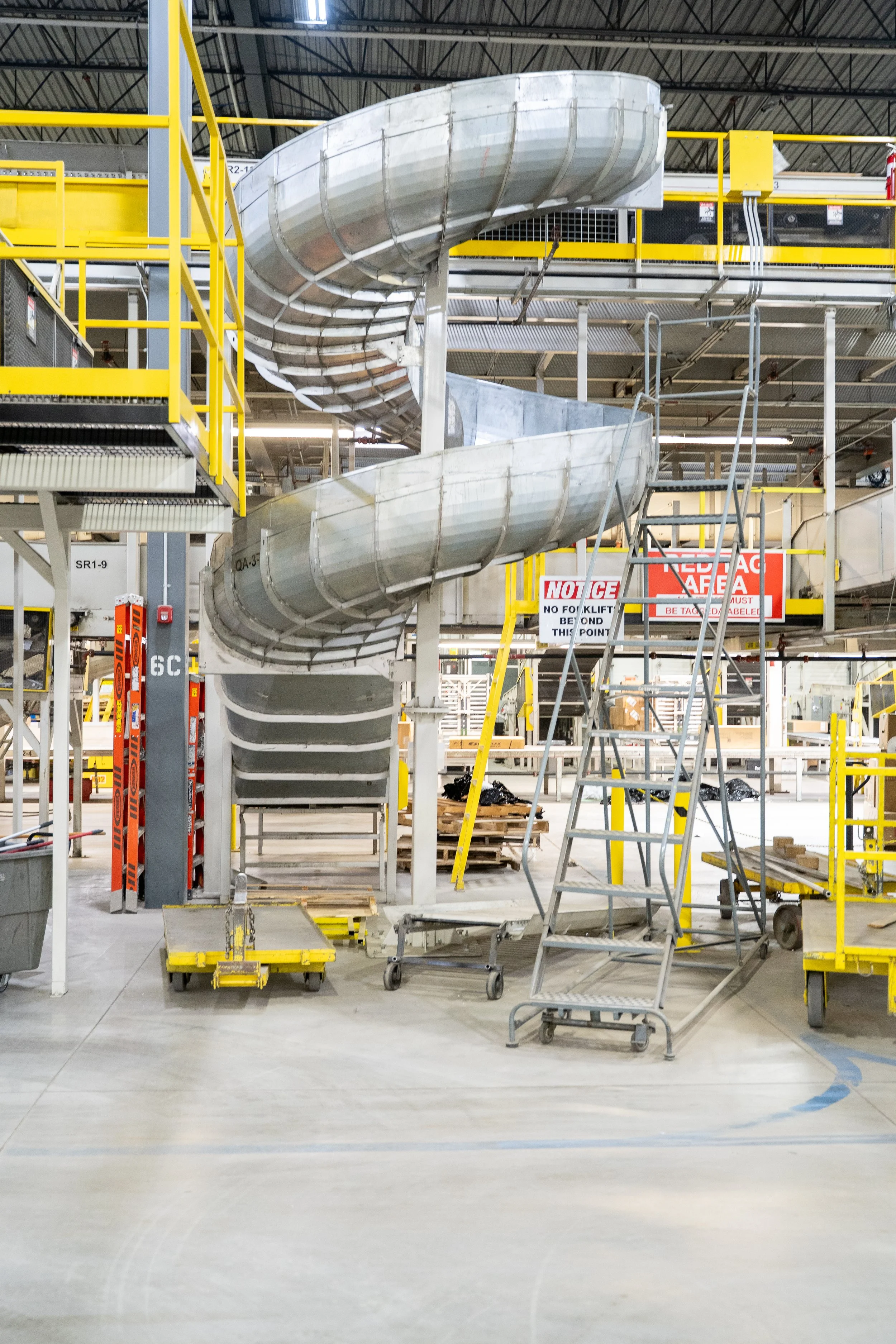Indoor industrial workspace with silver spiral slide, yellow safety rails, and ladders, surrounded by construction equipment and safety signs.