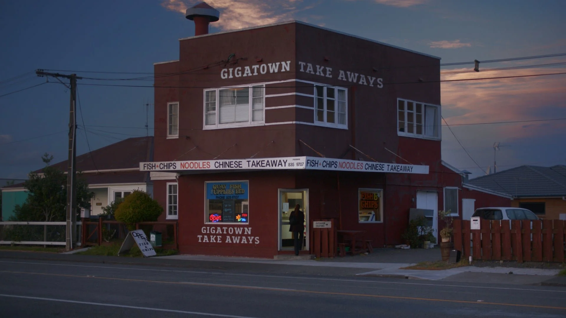 A two-story red building with signage reading 'GIGATOWN TAKE AWAYS,' offering fish, chips, noodles, and Chinese takeaway. An illuminated neon 'Open' sign is visible through the window, and a person is walking into the door during evening hours with a cloudy sky in the background.