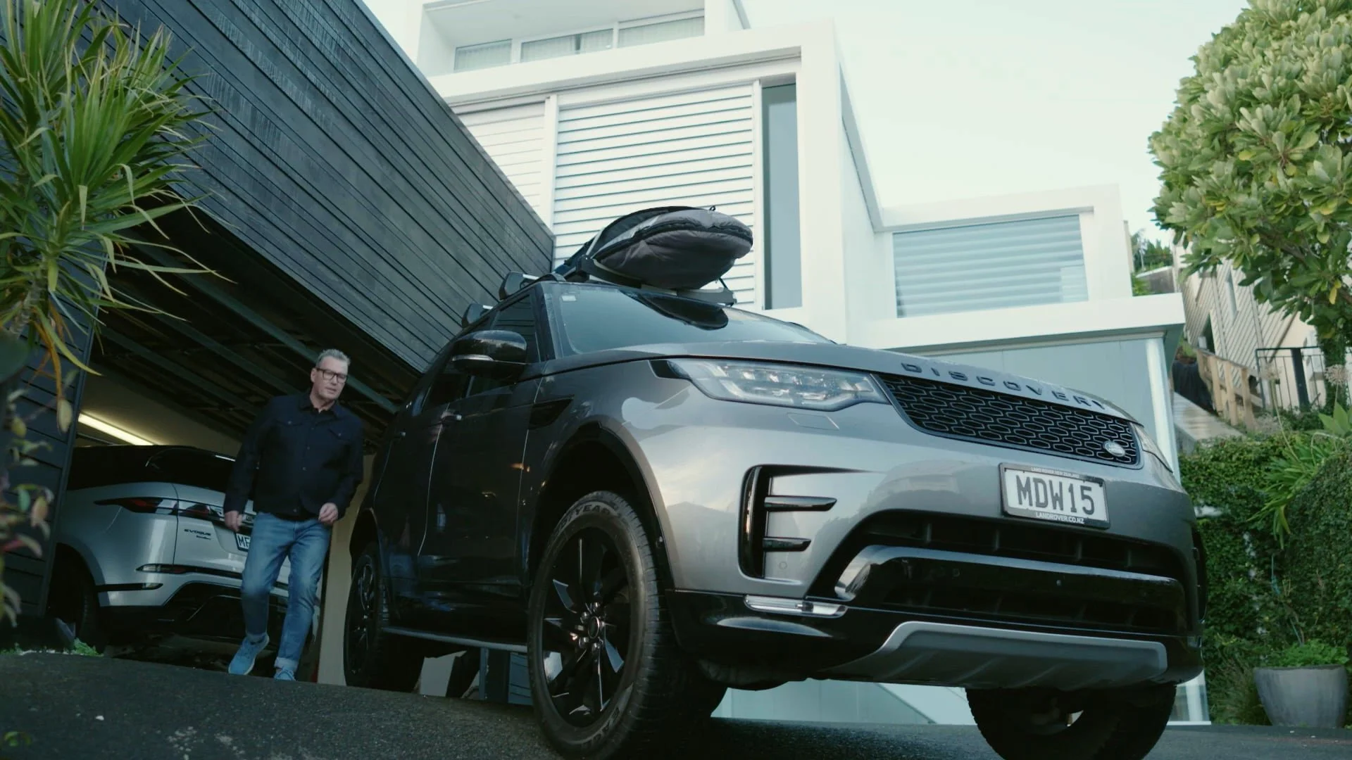 A man walking out of a modern house with a black and white Range Rover Sport parked in the driveway, with a roof box mounted on top of the vehicle.