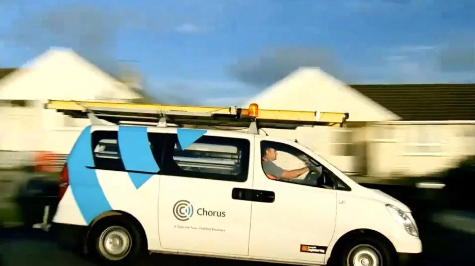 White utility van with 'Chorus' logo, moving at high speed, with a man driving, against a background of residential houses and a cloudy blue sky.