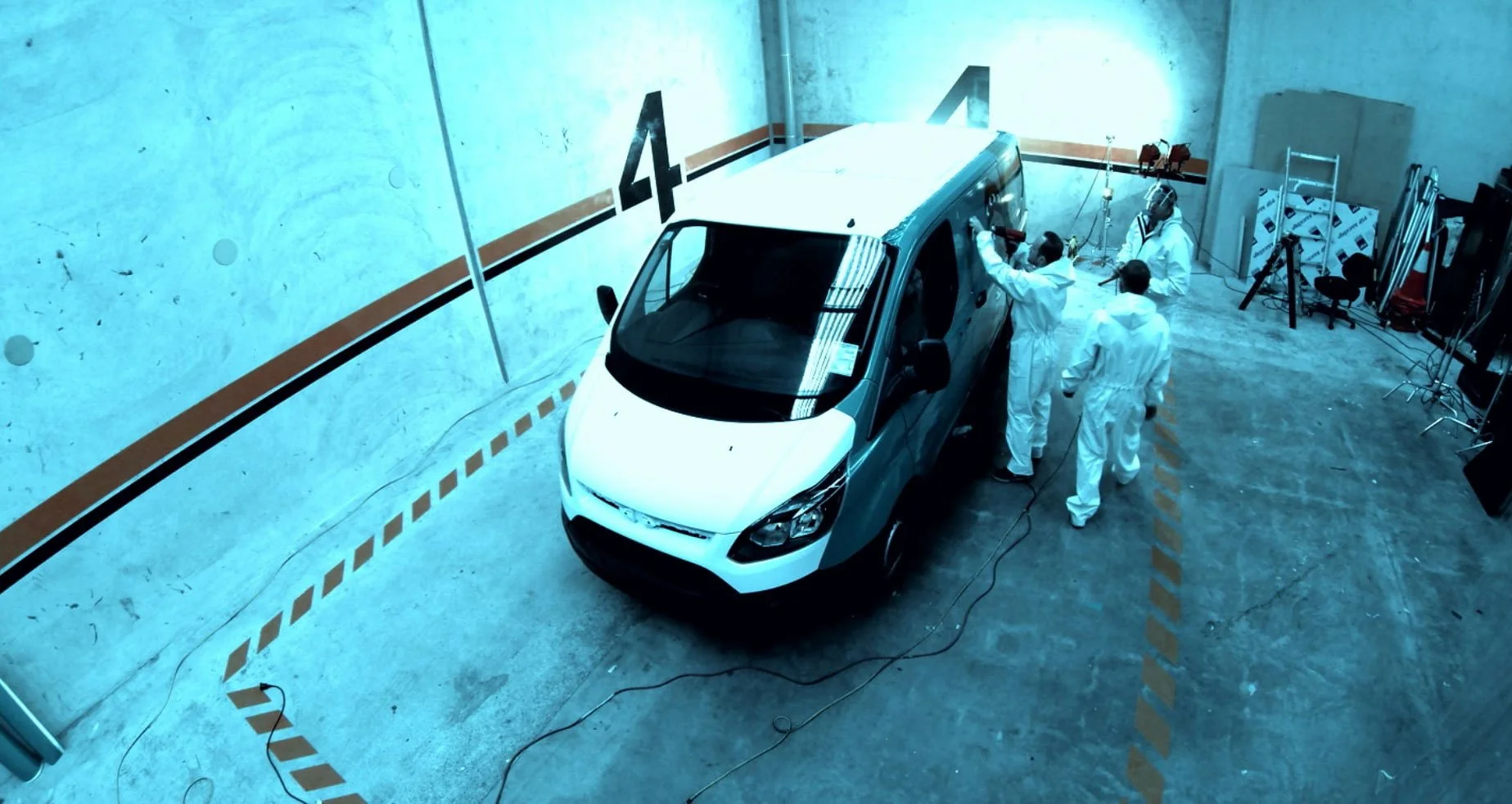 Four individuals in white coveralls working on a white car in a testing laboratory or wind tunnel with blue lighting, enclosed by white walls marked with black numbers 4 and 5.