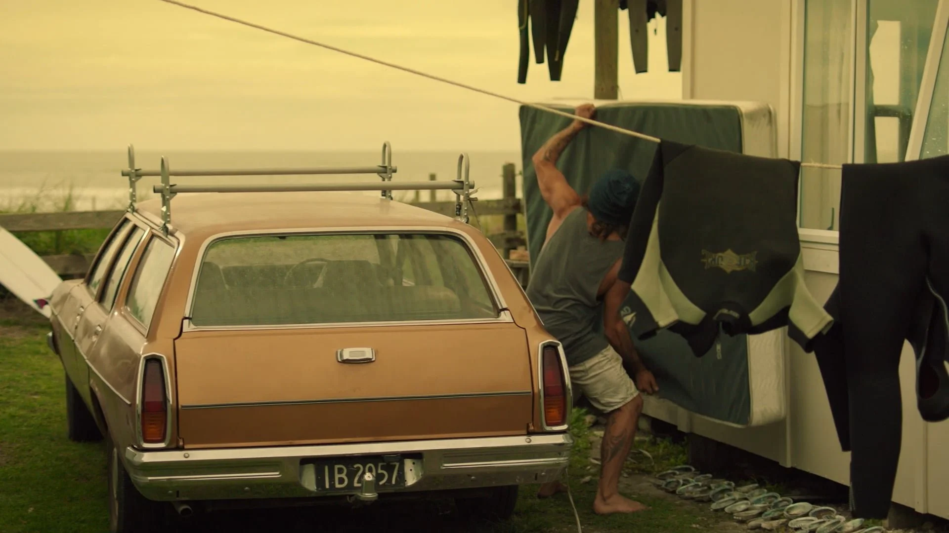 A man with long hair, wearing a beanie, is hanging laundry on a clothesline next to a vintage brown station wagon car parked on grass, with slippers lined up on the ground nearby.