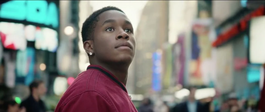 A young man with dark skin wearing a red jacket, standing in a busy urban area with blurred city lights and screens in the background.