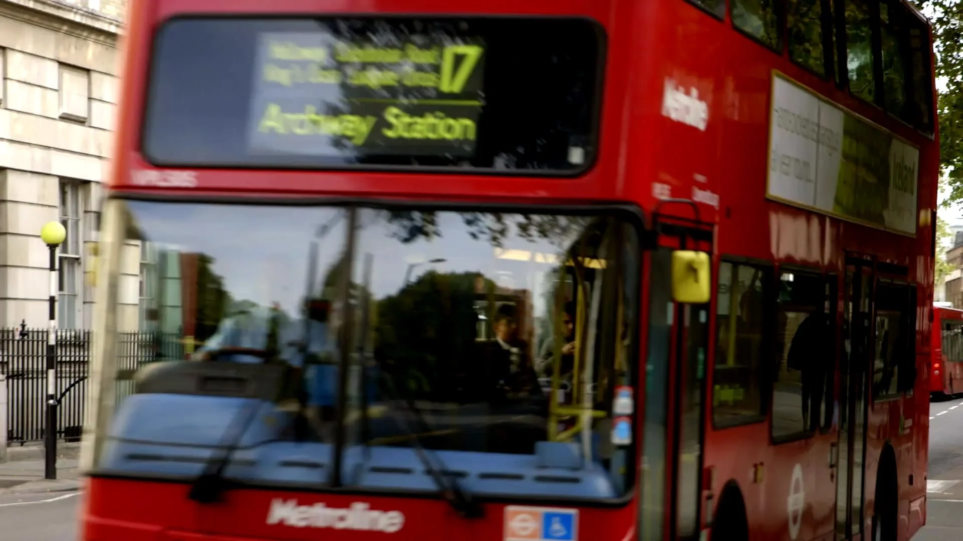Red double-decker bus on a city street, with an electronic sign displaying route information, buildings, and a black fence visible in the background.