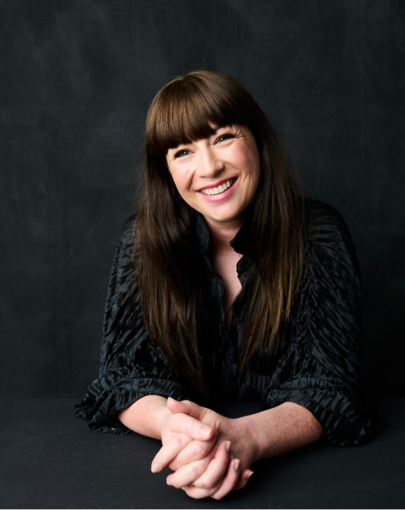 Smiling person with long brown hair wearing a black top, sitting with hands clasped on a dark surface against a dark background.