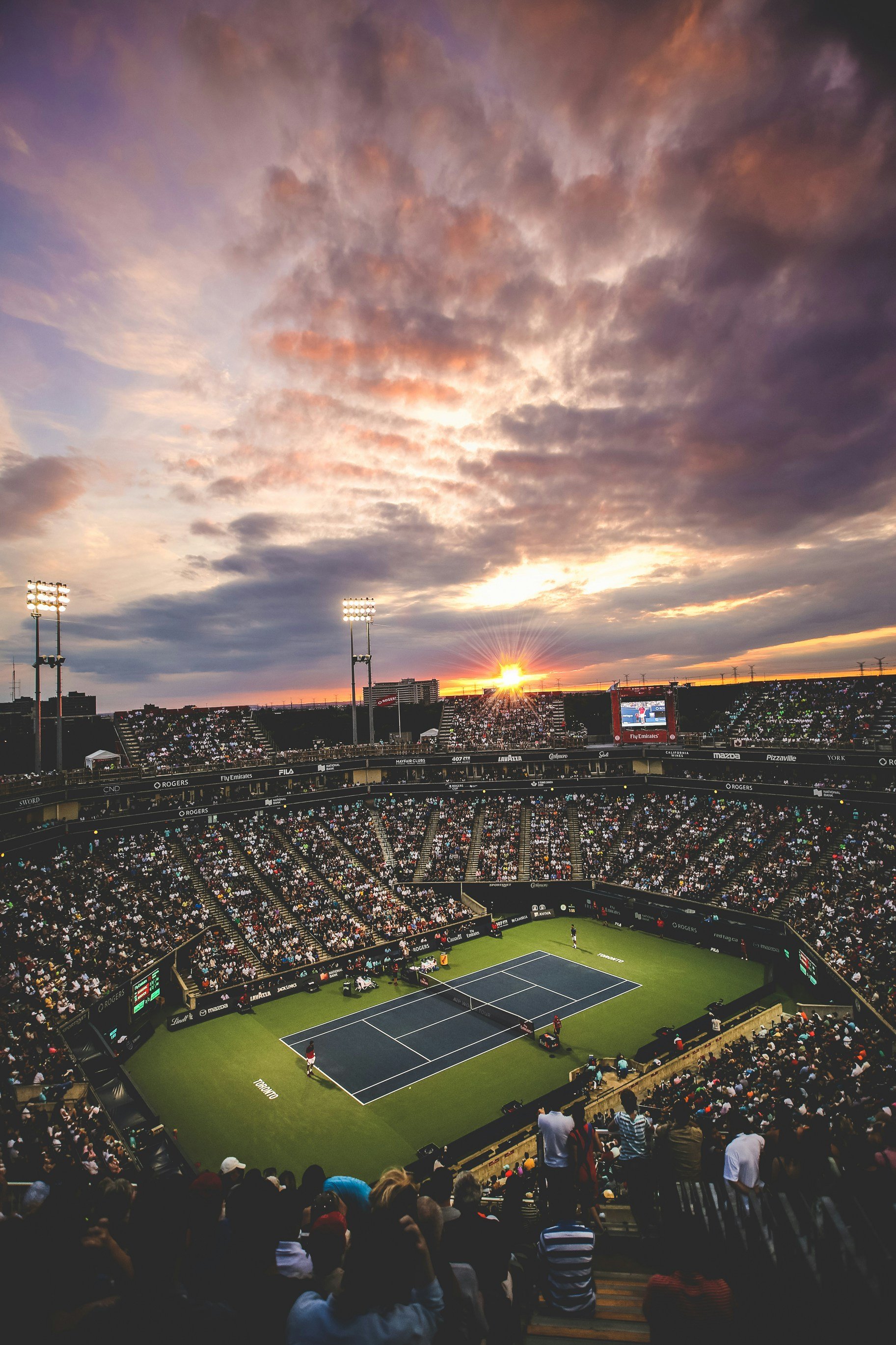 Tennis match in a stadium at sunset with a large crowd and dramatic clouds.