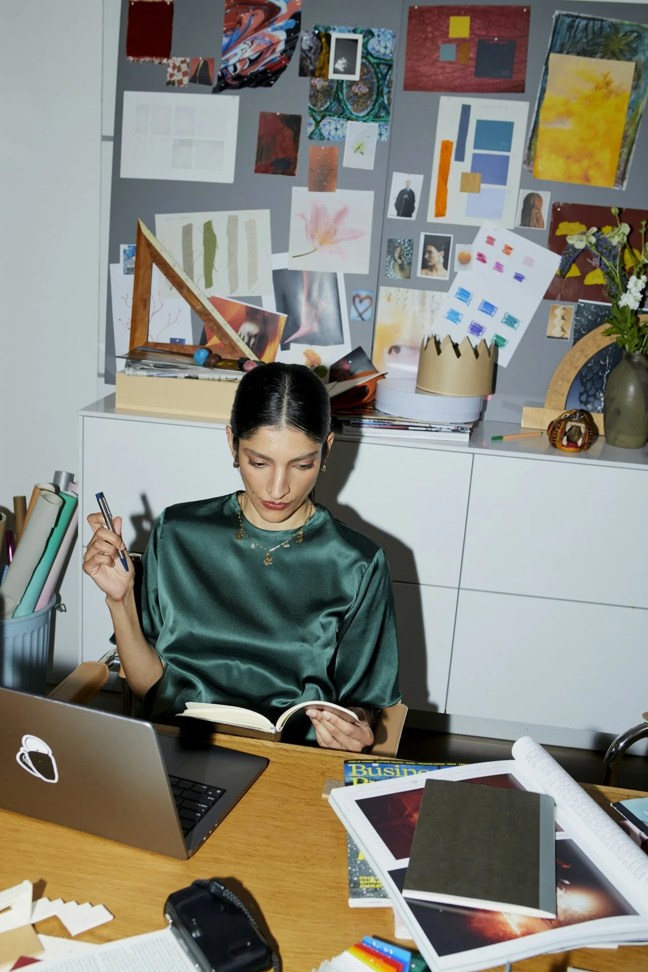 Woman in green satin blouse reading a book at a cluttered desk, surrounded by art supplies and a laptop, with a pinboard covered in colorful images and sketches in the background.