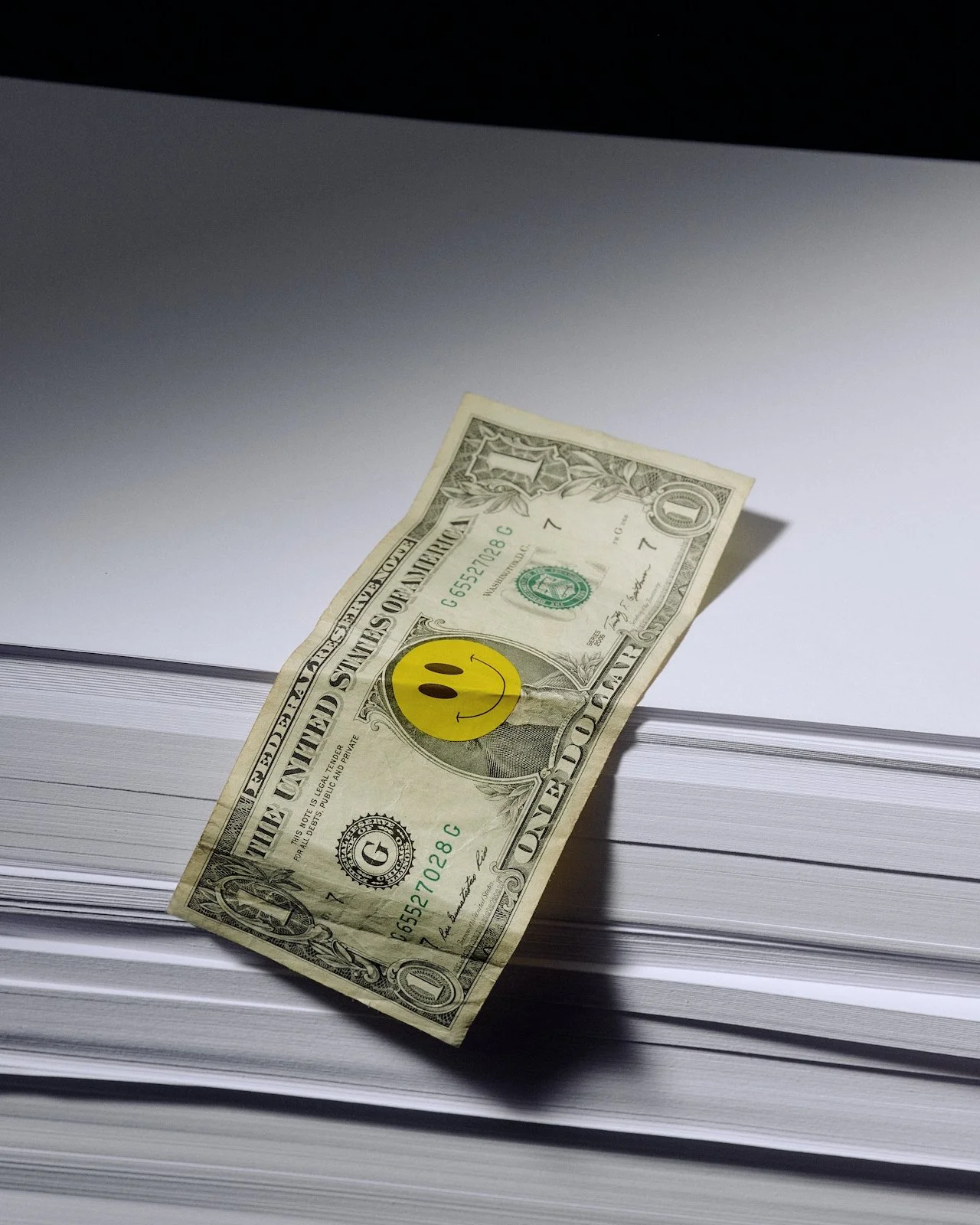 One US dollar bill with a yellow smiley face sticker on a stack of white paper.