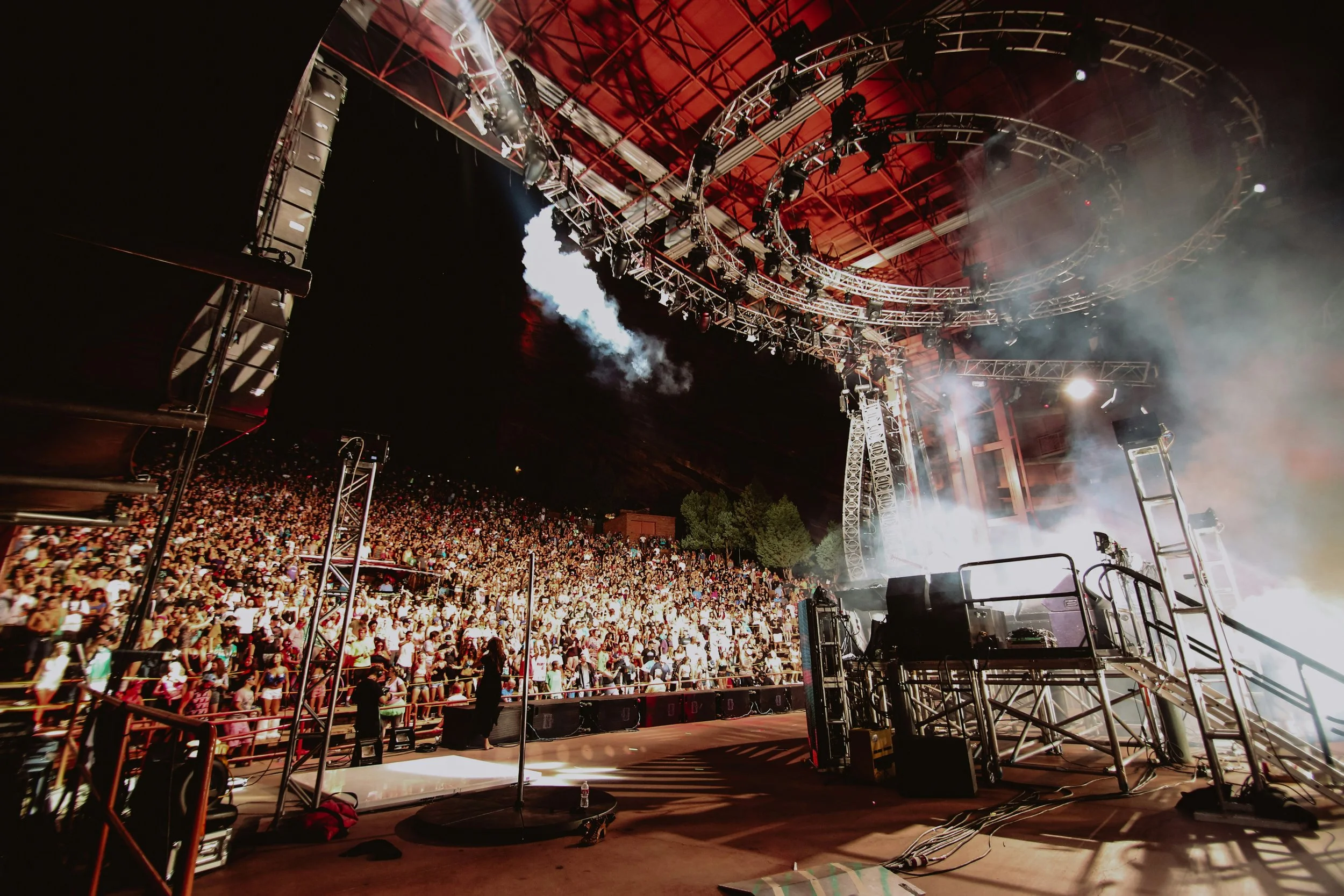Outdoor concert venue with large crowd, stage lighting, and smoke effects at night.