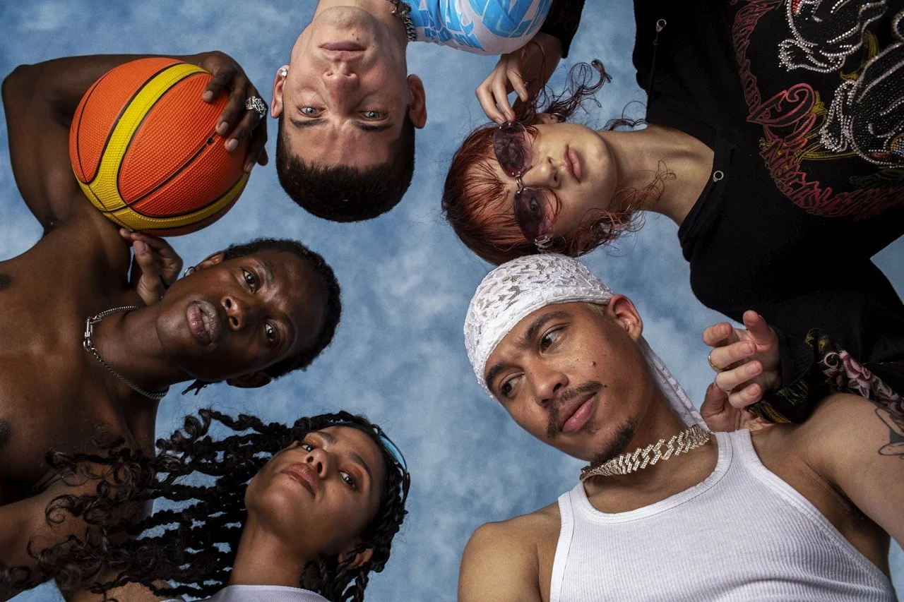 Five diverse people looking down at the camera, one holding a basketball, against a blue sky background.