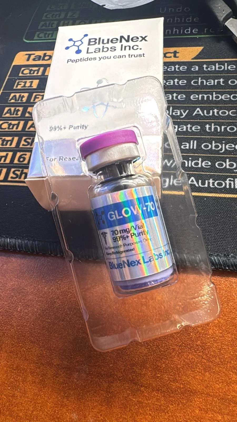 A small vial with a holographic label that reads 'GLOW-70, 70 mg/ vial, 99% Purity' from BlueNex Labs Inc., and a pre-filled syringe with a purple cap, placed inside clear plastic packaging on a black surface with printed instructions.