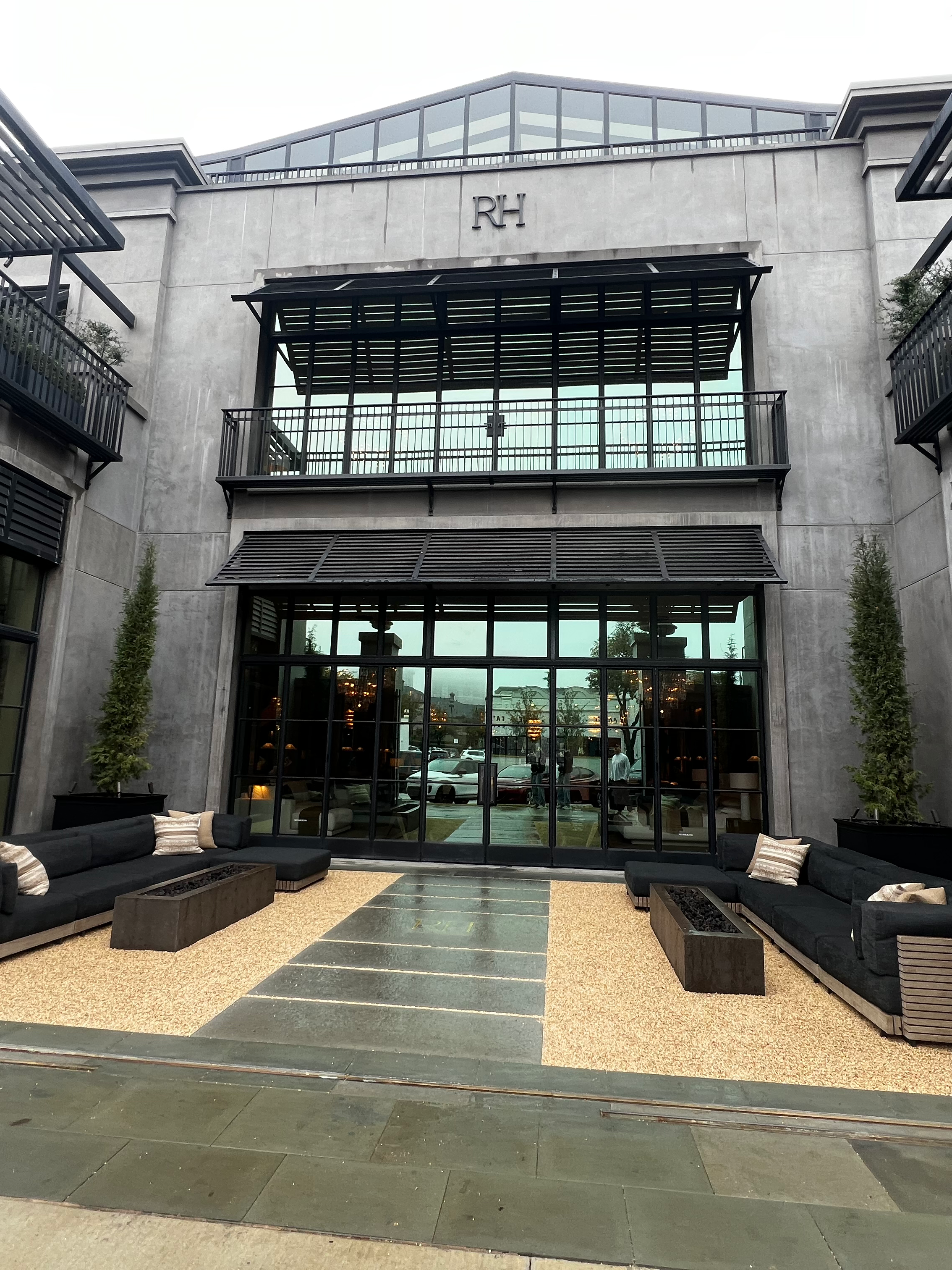 Modern multi-story building with a concrete facade, large glass windows, and balconies with black railings, labeled 'RH'. Outdoor seating area with black sofas and rectangular planters, on a mix of gravel and stone surface, with trees and parking lot visible in the background.