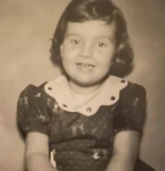 A young girl with short, curly hair smiling for a portrait photograph, wearing a dark dress with a light-colored collar.