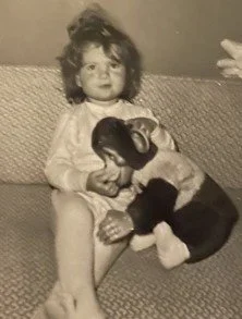A young girl with curly hair sitting on a couch, holding a plush toy dog while cuddling a small puppy.