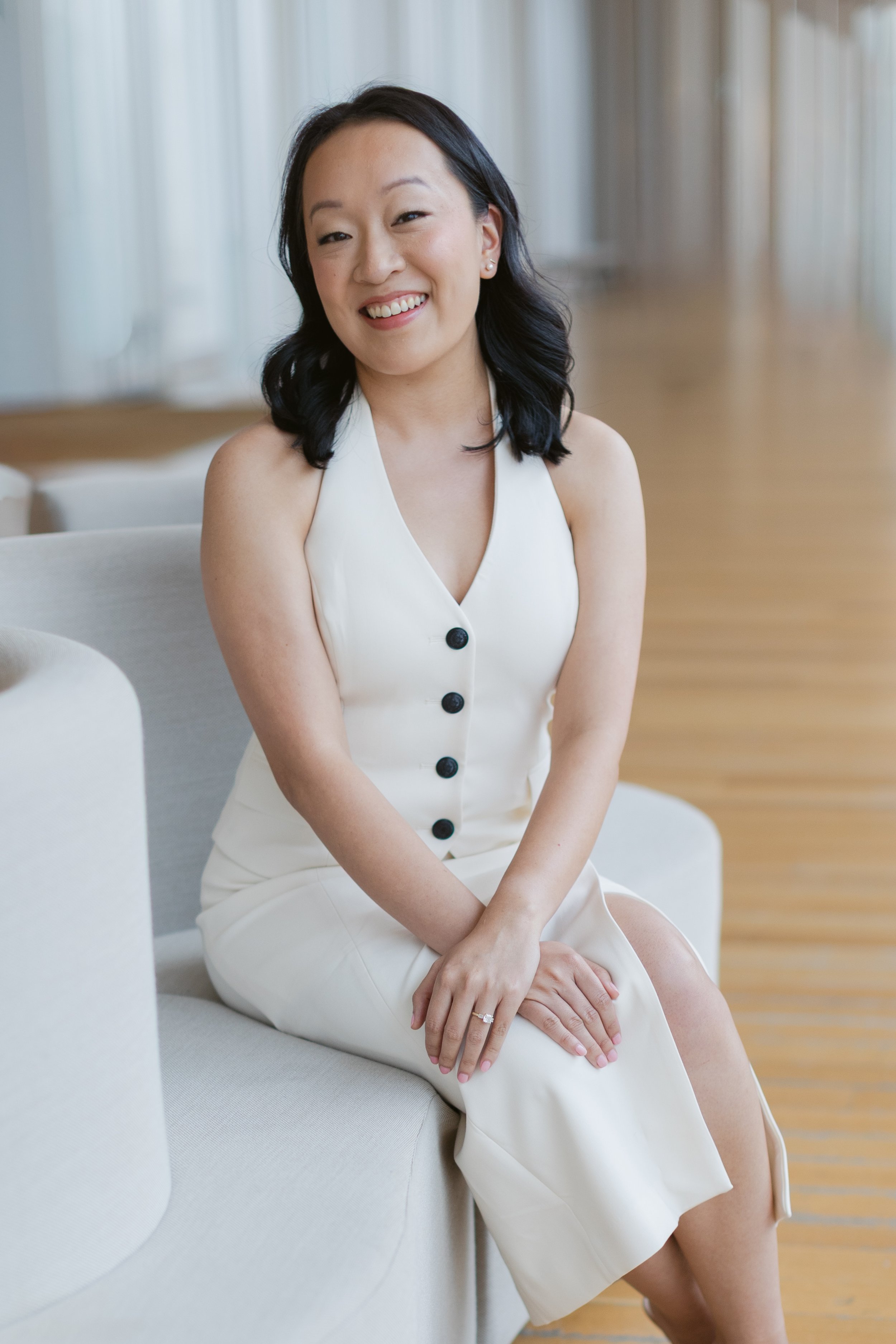 A woman with black hair and light skin tone smiling, sitting on a cream-colored sofa in a bright room with large windows, wearing a white sleeveless dress with black buttons.