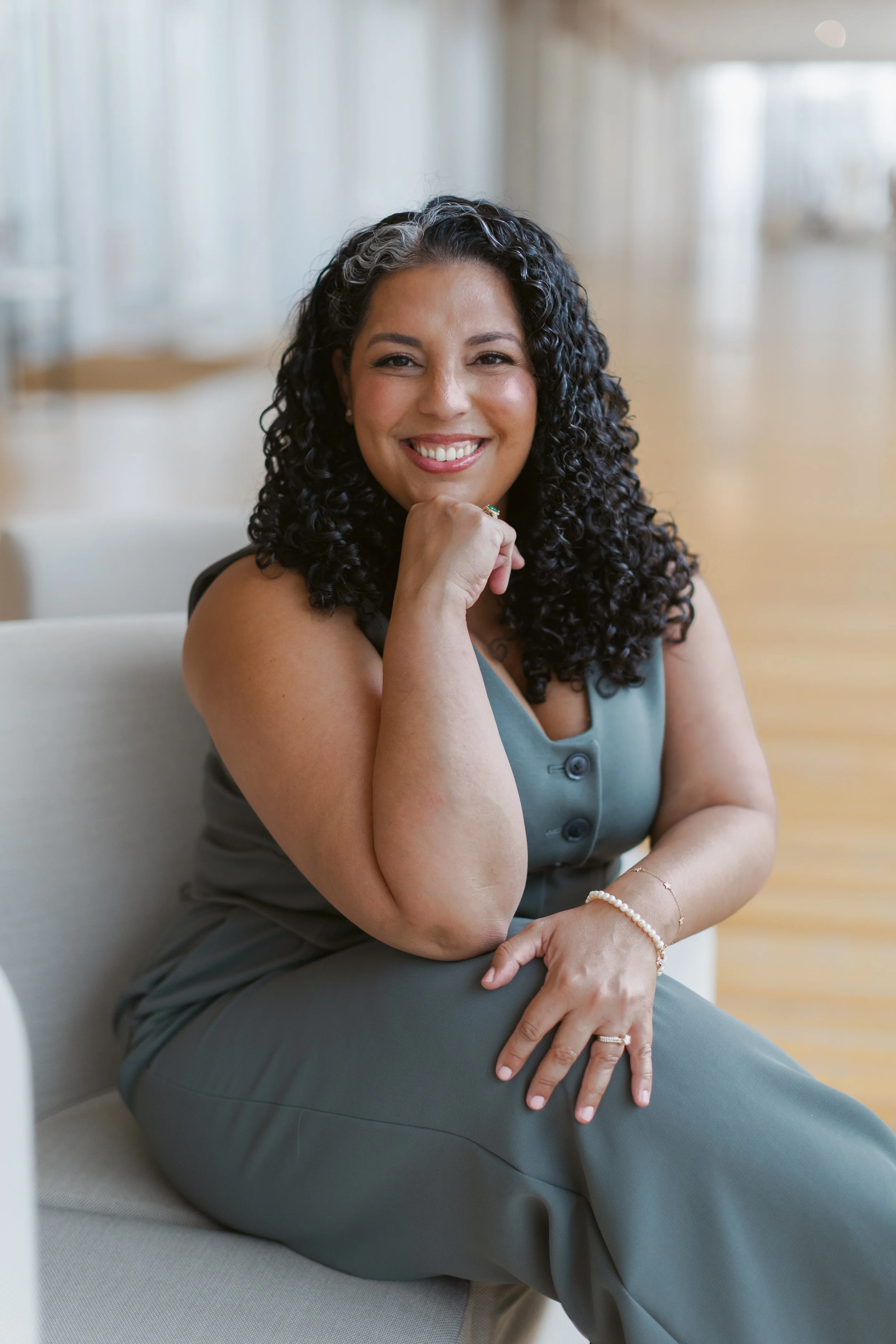 A woman with dark, curly hair smiling and sitting on a beige chair in a well-lit room, wearing a sleeveless green pant suit and jewelry.