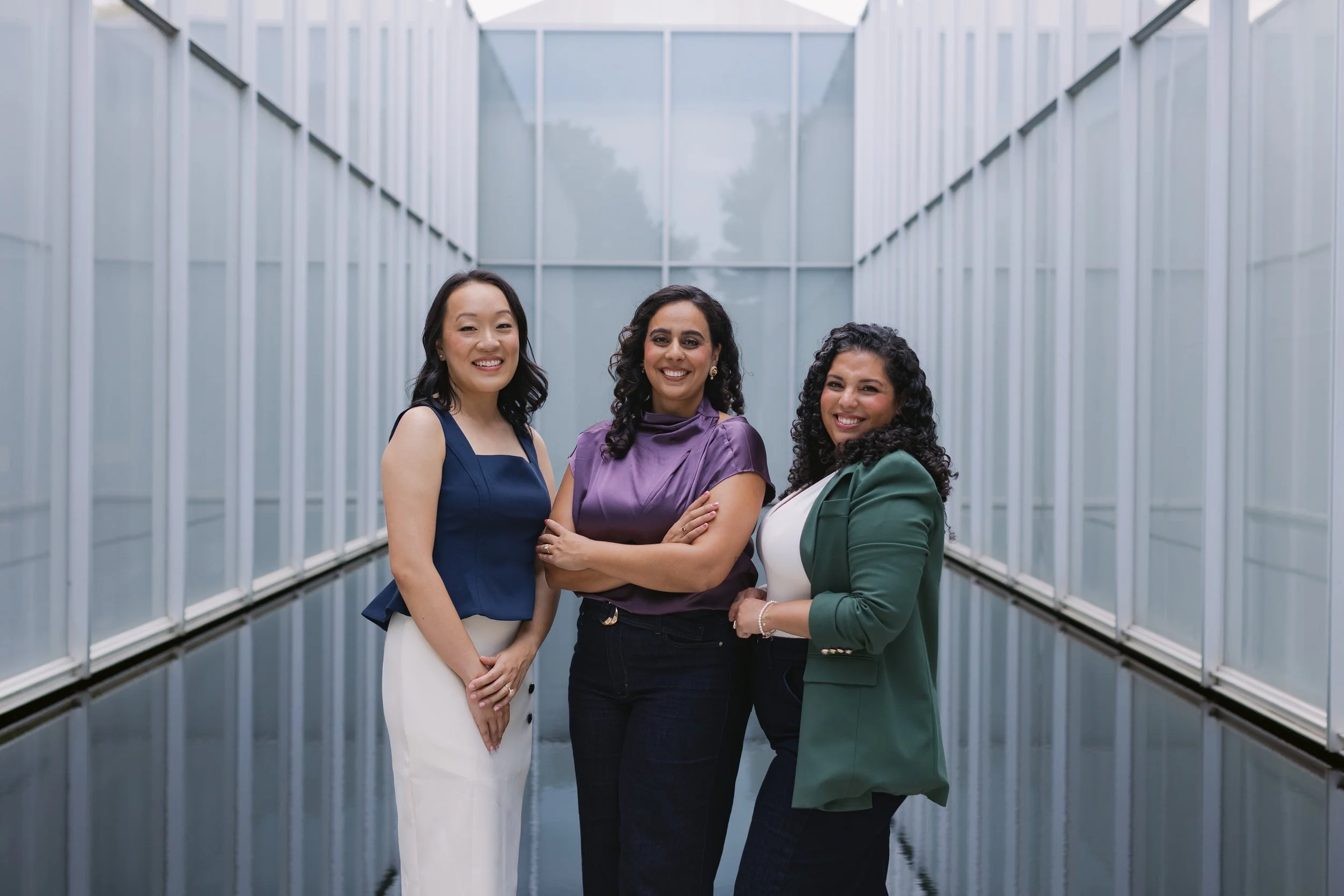 Three women standing together inside a glass corridor, smiling at the camera.