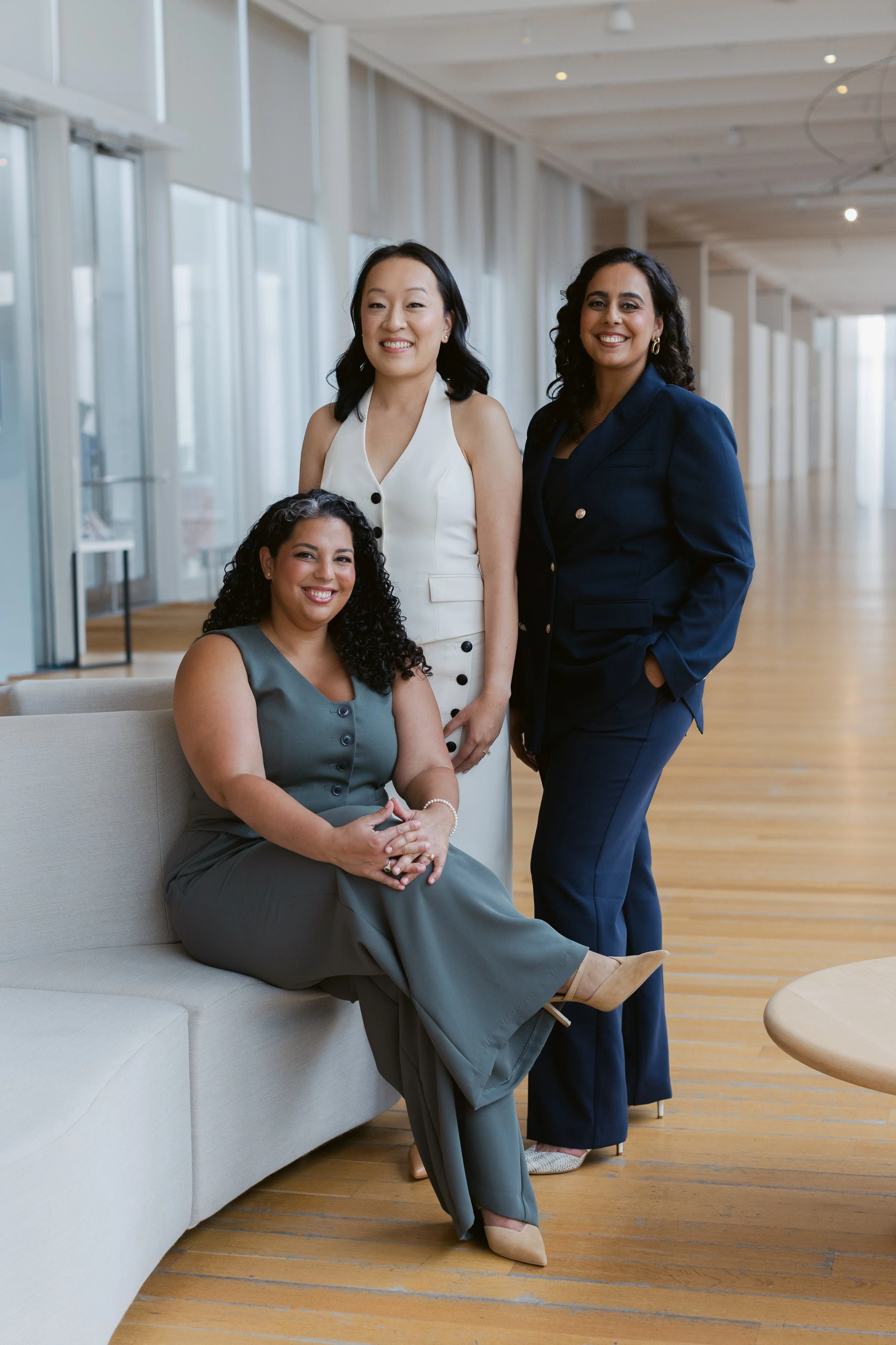 Three professional women in business attire, smiling, in a modern indoor space with large windows and wooden floors.
