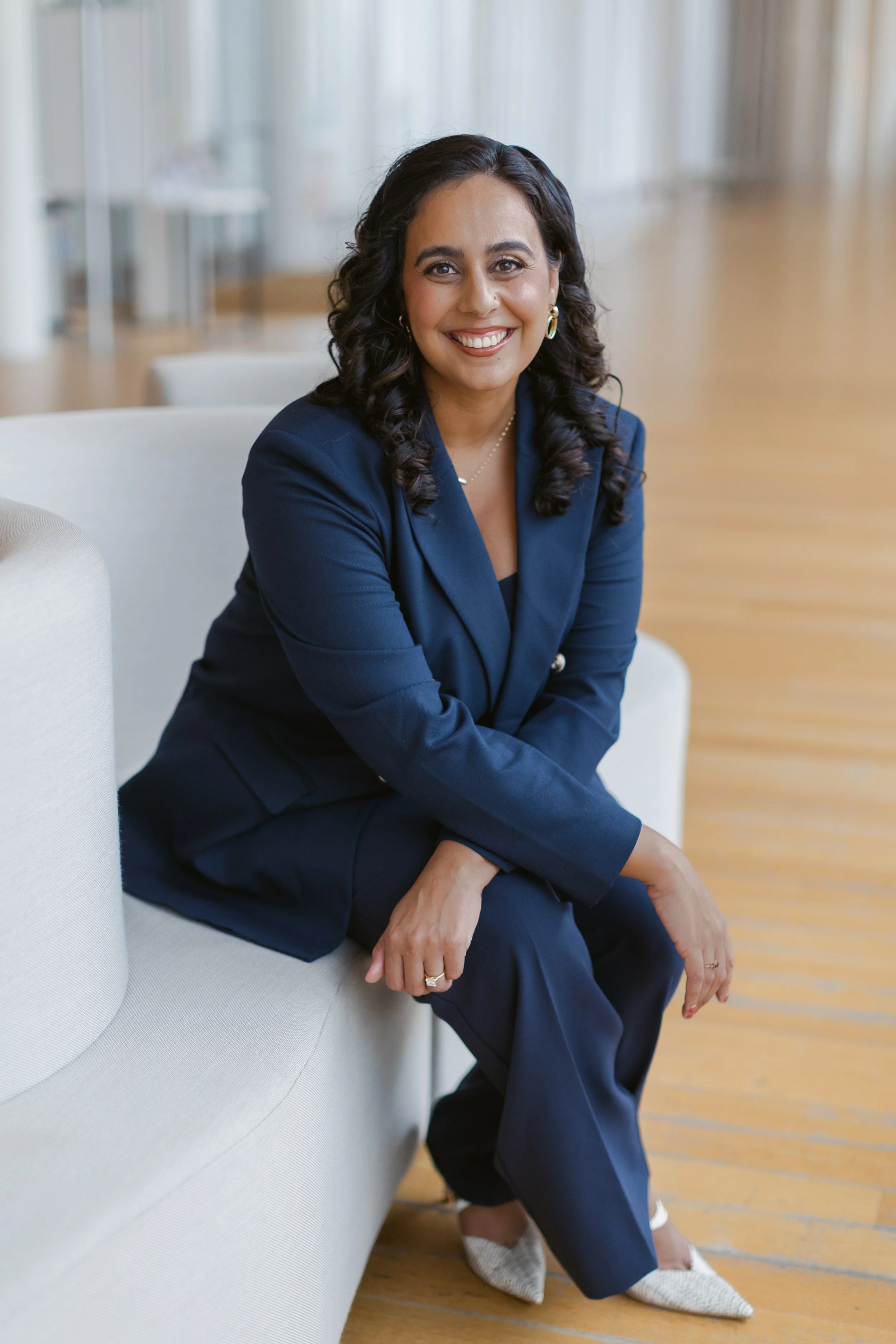 A woman with dark curly hair, wearing a navy blazer and navy pants, sitting on a white sofa in a bright, modern room with wooden floors.