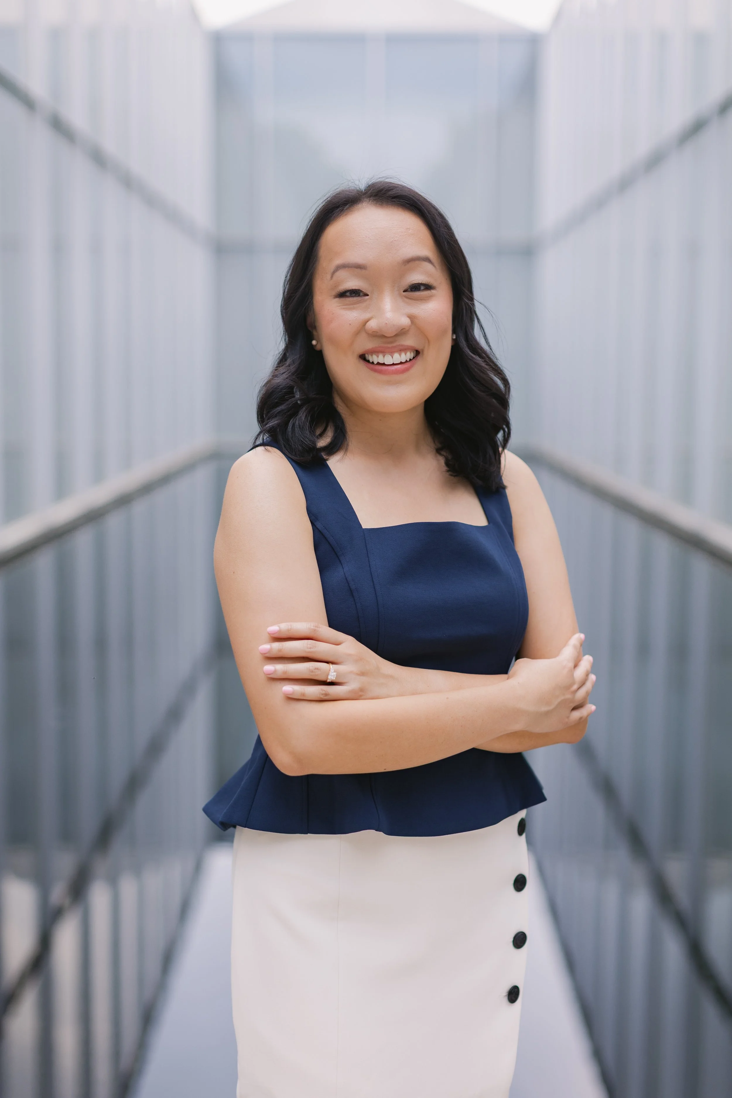 A woman in a navy blue sleeveless top and white skirt standing in a metal walkway, smiling with arms crossed.