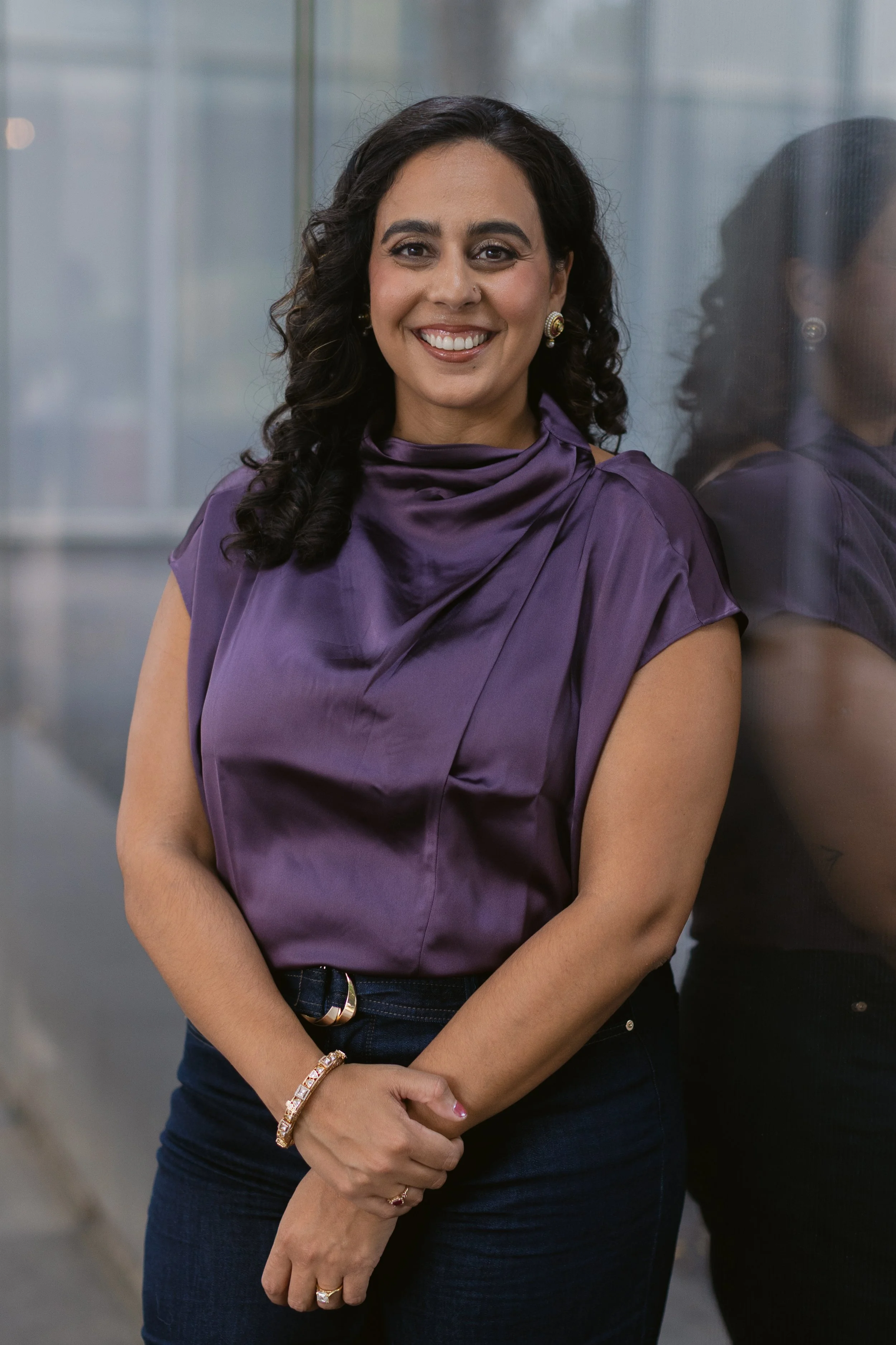 Woman with dark curly hair smiling, wearing a purple satin top and dark jeans, standing indoors near a glass wall.