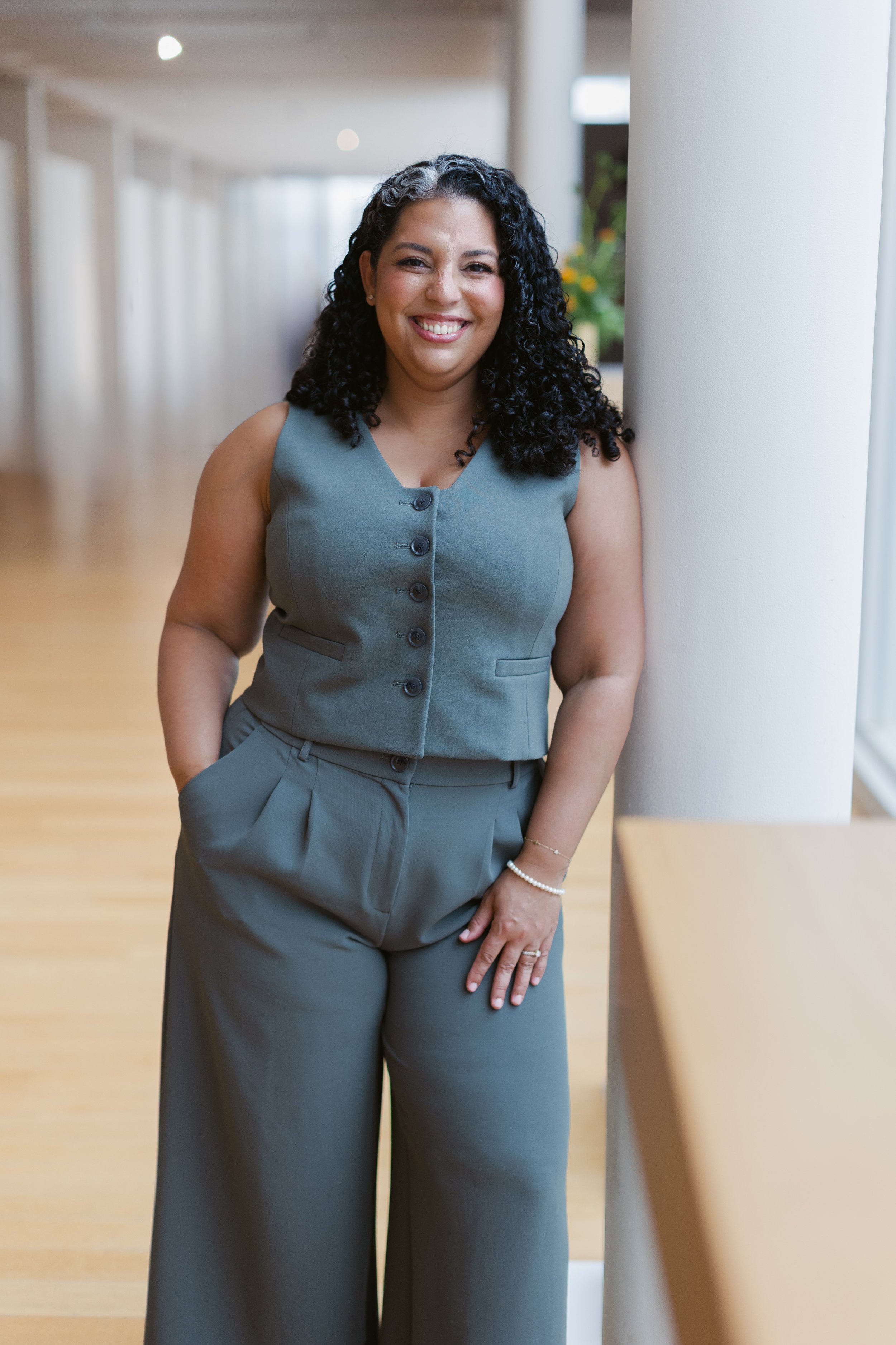Smiling woman in a bluish-gray sleeveless suit, standing indoors near a white pillar, with her hands in her pockets, in a modern building with wooden flooring and natural light.